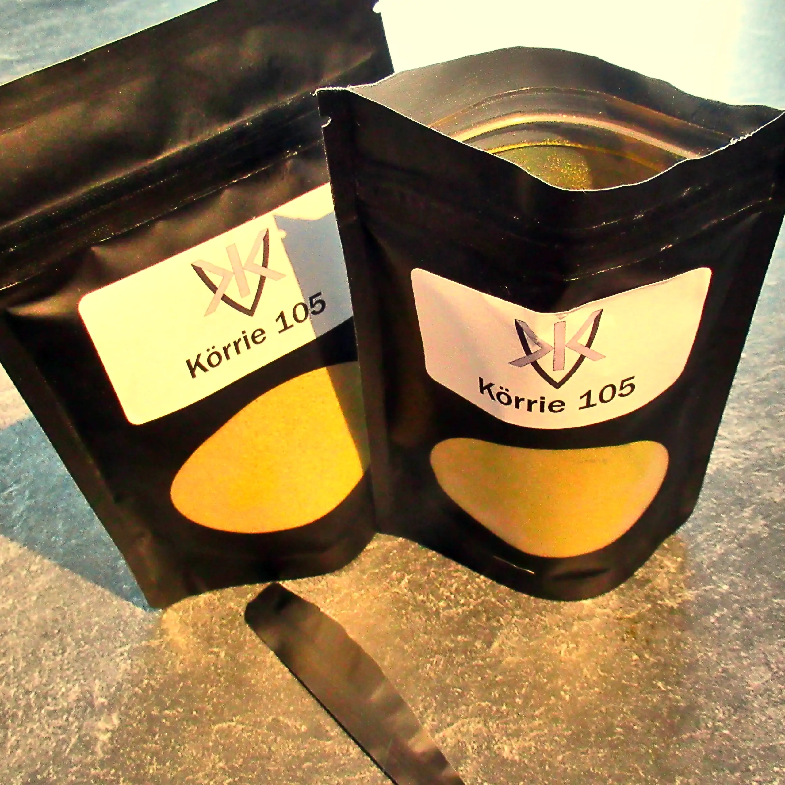 Ergänzende Ansicht der Körrie 105 Gewürzmischung: Zwei schwarze Beutel mit transparentem Sichtfenster, durch das die leuchtend goldgelbe, fein gemahlene Currypulver-Mischung sichtbar ist, versehen mit weißen Labels, die das Logo und "Körrie 105" zeigen (e