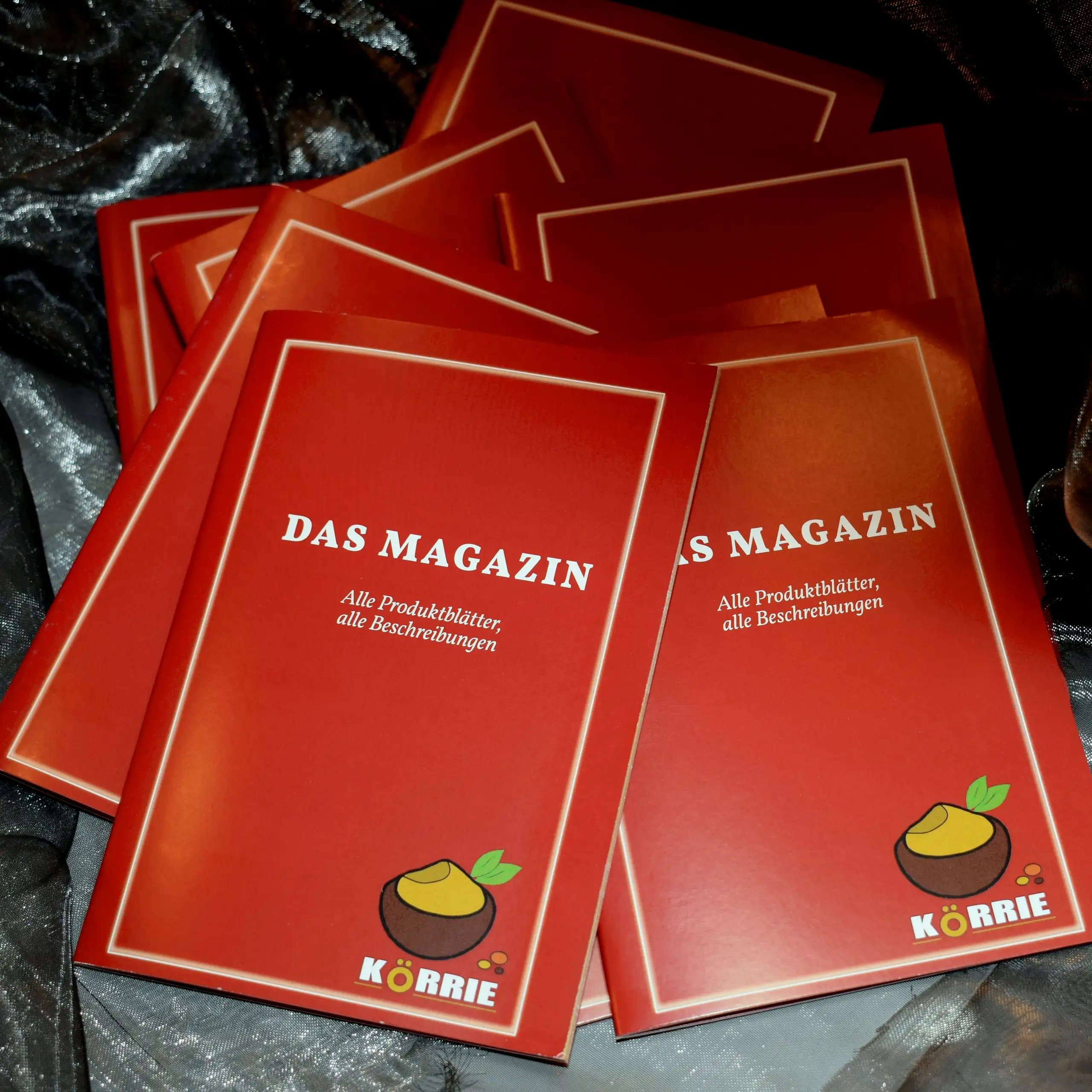 Ergänzende Ansicht des Körrie-Support-Magazins / Körrie KSM: Ein Stapel roter Magazine mit dem Körrie-Logo, dem Titel "DAS MAGAZIN" und Untertitel "Alle Produktblätter, alle Beschreibungen" in weißer Schrift, präsentiert auf einem dunklen, schimmernden Hi