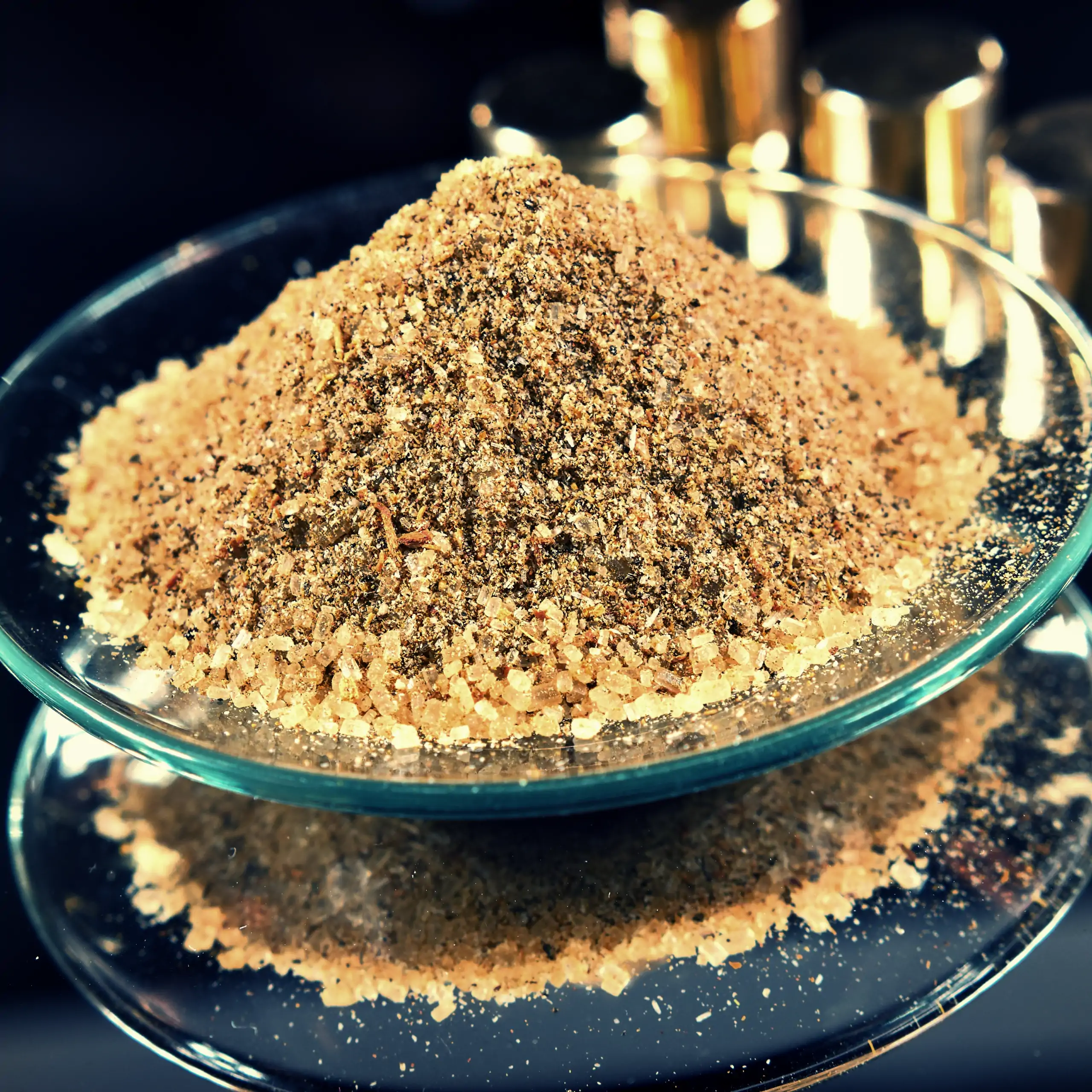 Detailaufnahme der Körrie WVZ Mischung: Ein Haufen leuchtend goldbrauner, fein gemahlener würzig-süßer mit deutlich erkennbaren Kristallen von Rohrzucker auf einer transparenten Glasschale auf dunklem Hintergrund mit goldenen reflektierenden Dekorationsel