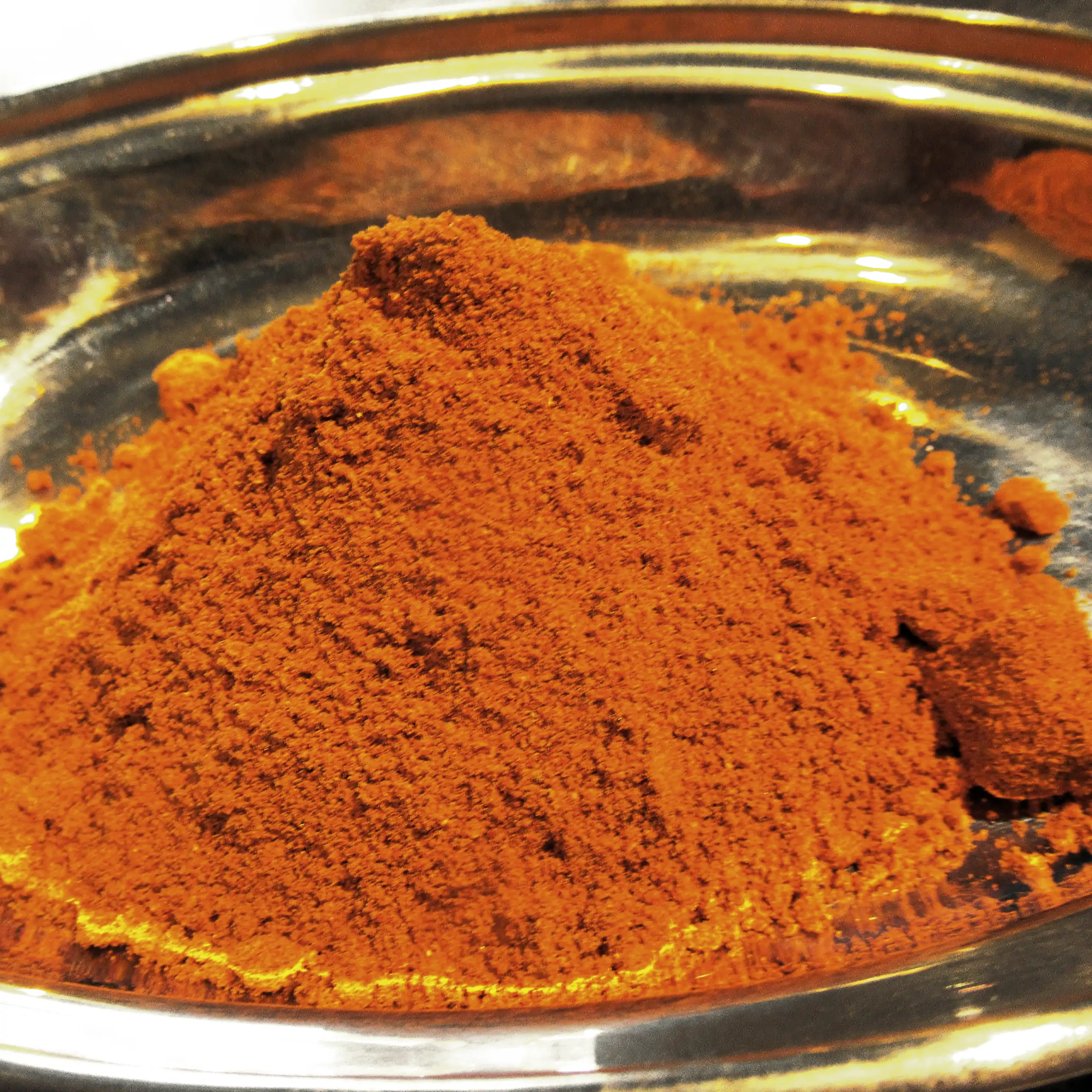 Ergänzende Ansicht der Körrie H55 Gewürzmischung: Ein Haufen leuchtend orangene-roter, fein gemahlener, herzhafter Gewürzmischung in einer metallischen Schale auf dunklem Hintergrund, der die intensive, aromatische Textur und vanillige Nuance betont, für 