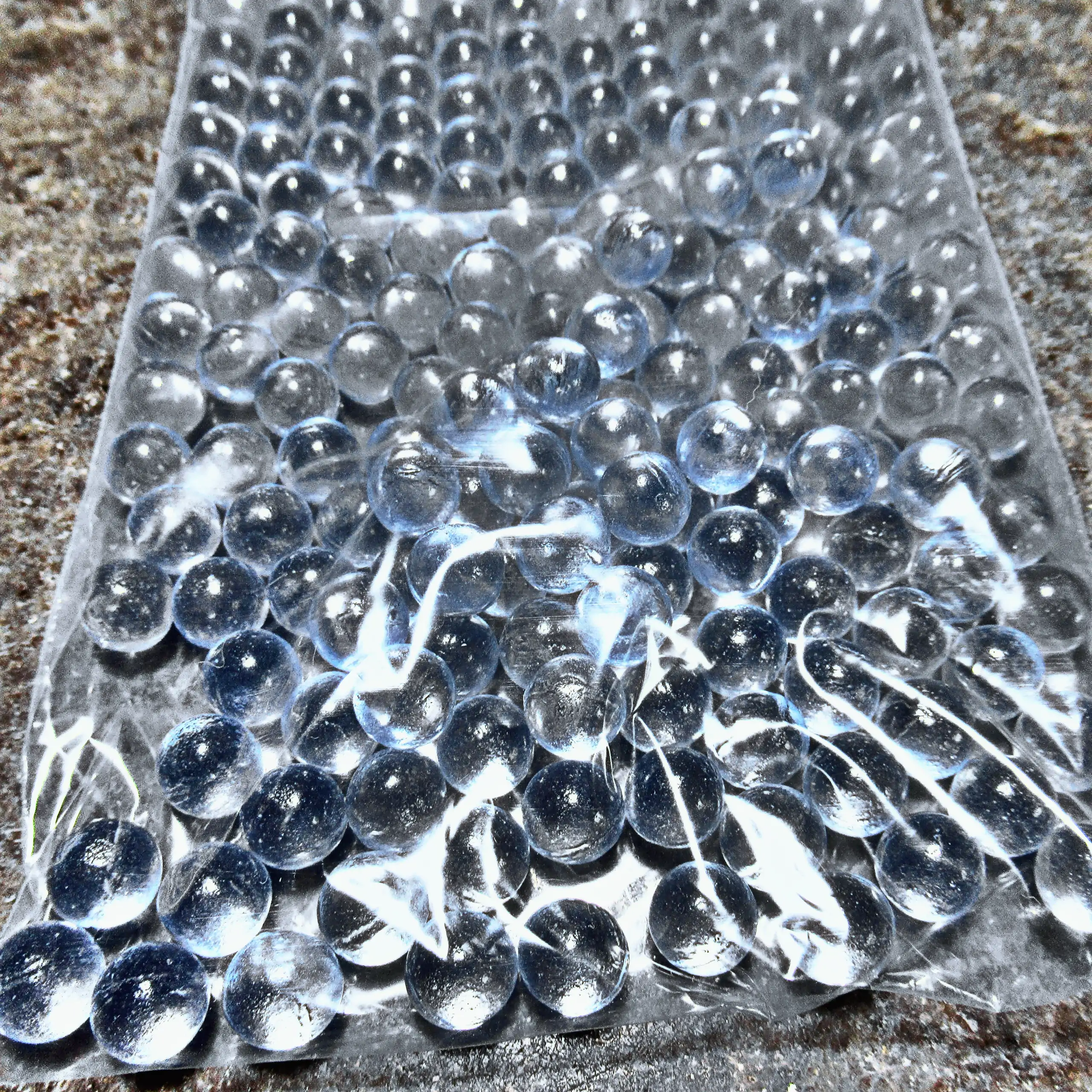 Zusätzliches Bild der Körrie-Aktiv-Perlen (Glas) / Körrie KAP L: Eine große Menge leuchtend transparenter, glänzender, gehärteter Glasperlen in einer durchsichtigen Plastikverpackung, verstreut auf einer dunklen Oberfläche mit funkelnden Lichtreflexen für