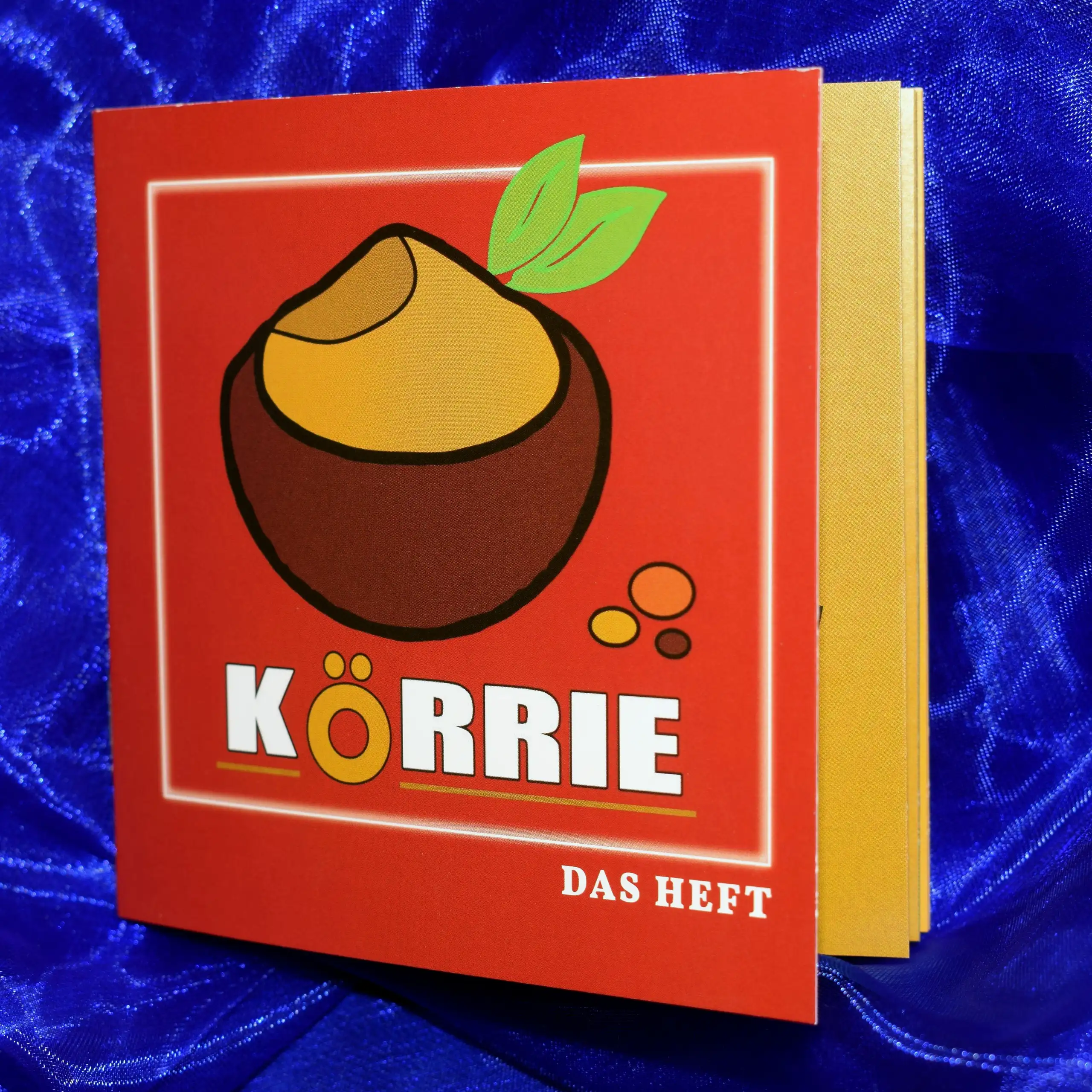 Detailaufnahme des Körrie-Support-Hefts / Körrie KSH: Ein leicht geöffnetes rotes Heft mit dem Körrie-Logo und dem Titel "KÖRRIE DAS HEFT" in weißer Schrift, präsentiert auf einem blauen Satin-Hintergrund für einen premium und einladenden Look. Dieses hoc