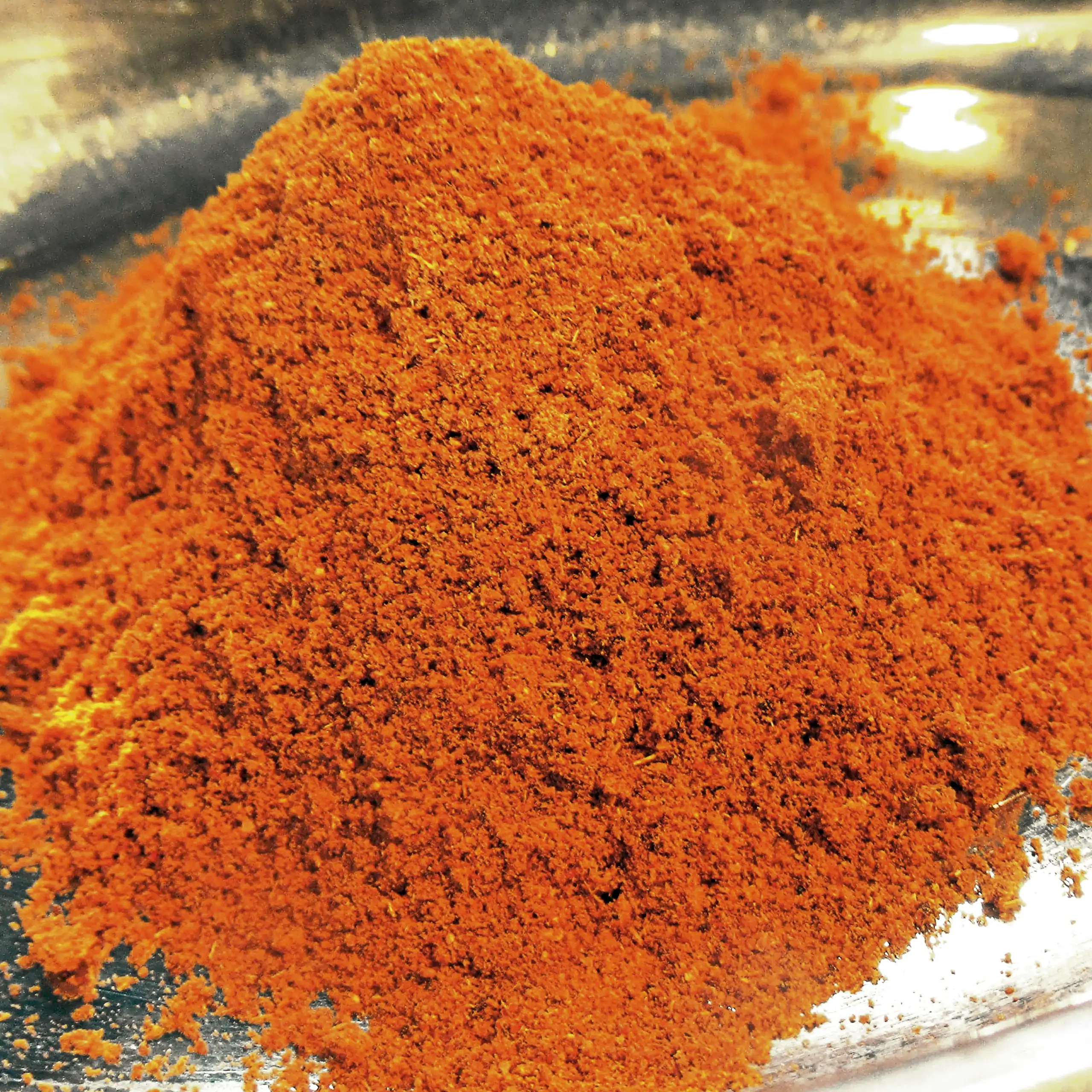 Ergänzende Ansicht der Körrie 011 Gewürzmischung: Ein Haufen leuchtend orange-roter, fein gemahlener rauchig-scharfer Curry-Gewürzmischung in einer flachen Edelstahlschale in Nahaufnahme, der die intensive, aromatische Textur und rotgoldene Nuance betont 