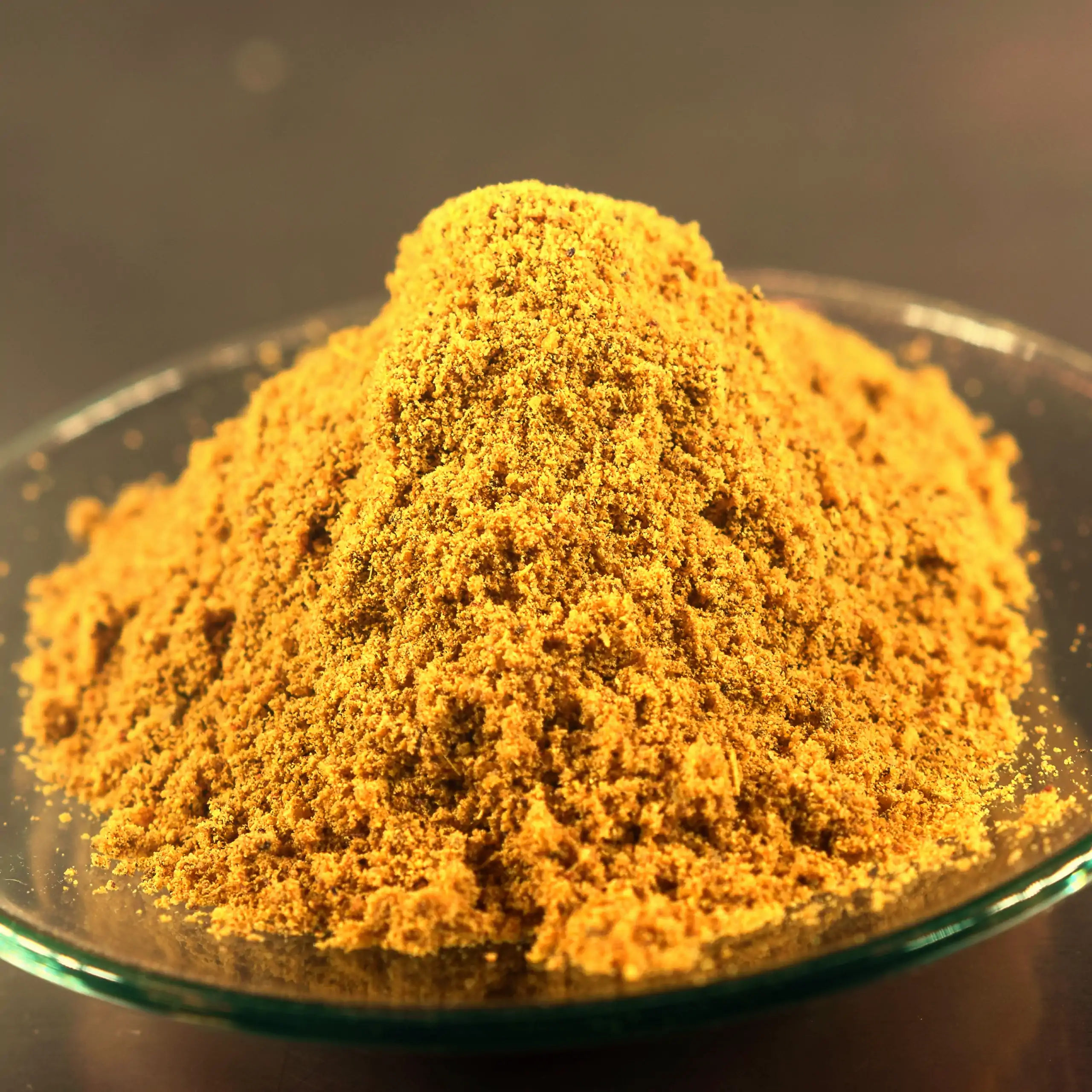 Detailaufnahme der Körrie 105 Gewürzmischung: Ein Haufen leuchtend goldgelber, fein gemahlener Currypulver-Mischung in einer transparenten Glasschale auf dunklem Hintergrund, der die intensive, aromatische Textur betont, für einen appetitlichen, natürlich