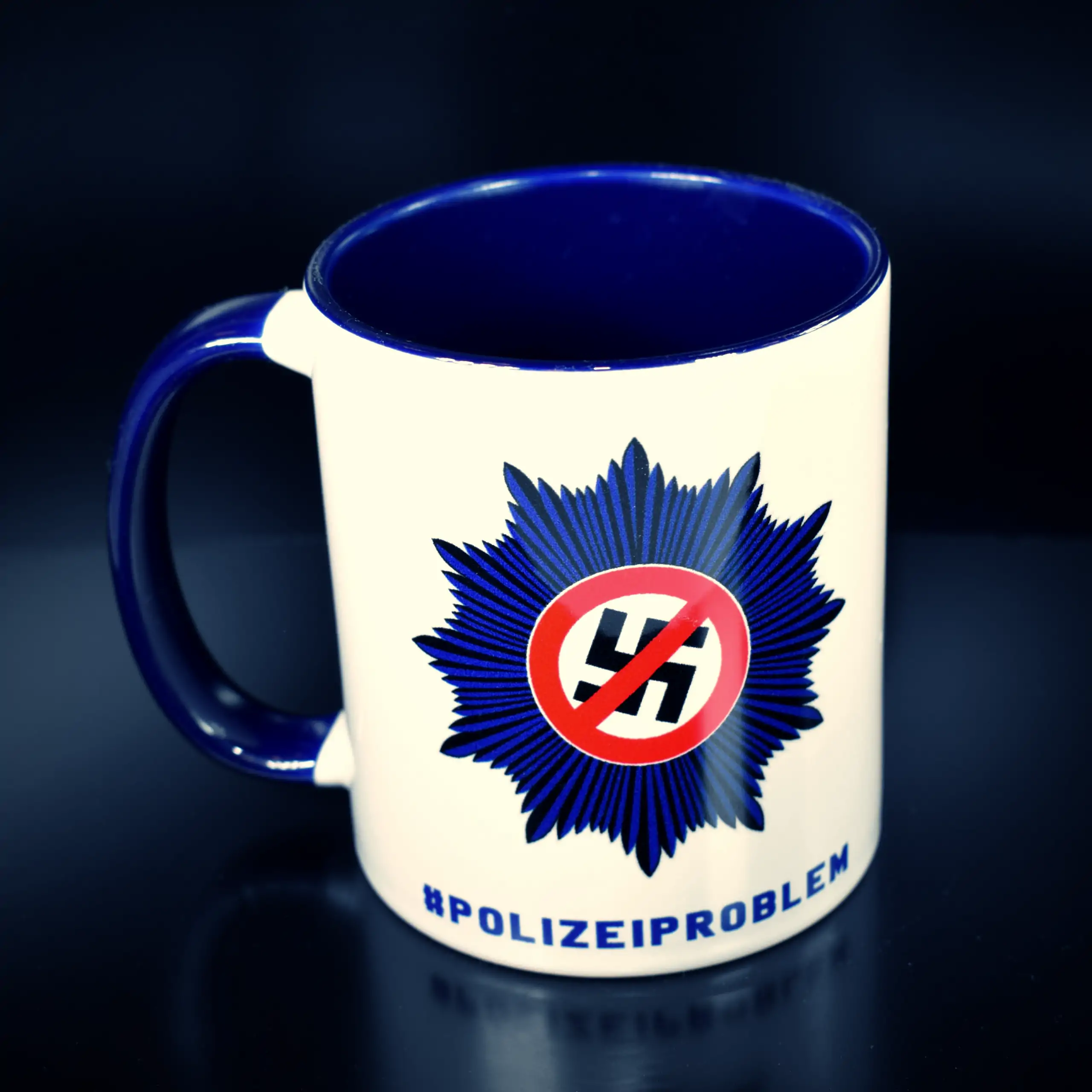 Titelbild der Tasse "Nazis raus aus der Polizei" (Variante: Griff links): Eine weiße Keramiktasse mit blauem Innenrand und Henkel, präsentiert auf einem dunklen Untergrund für einen kontrastreichen, markanten Look, wobei der Henkel links vom Logo position