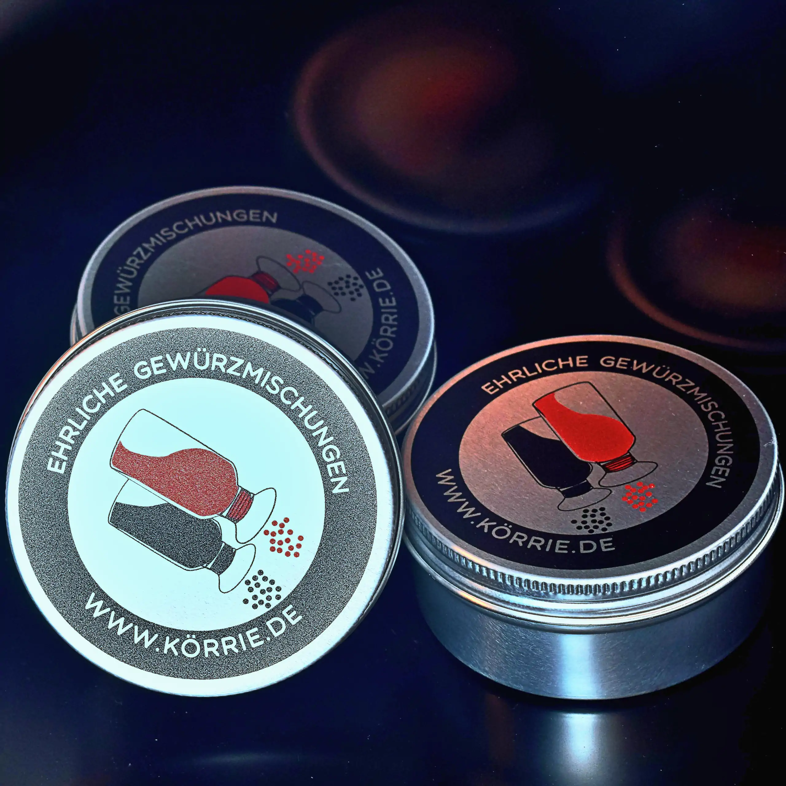 Ergänzende Ansicht der Körrie-Support-Dose: Drei kompakte, silberfarben glänzende Metall-Dosen mit schwarz-rot bedruckten Deckeln, offen präsentiert auf dunklem Hintergrund mit warmen Lichtreflexen im Hintergrund. Die Deckel tragen das Körrie-Logo mit umg