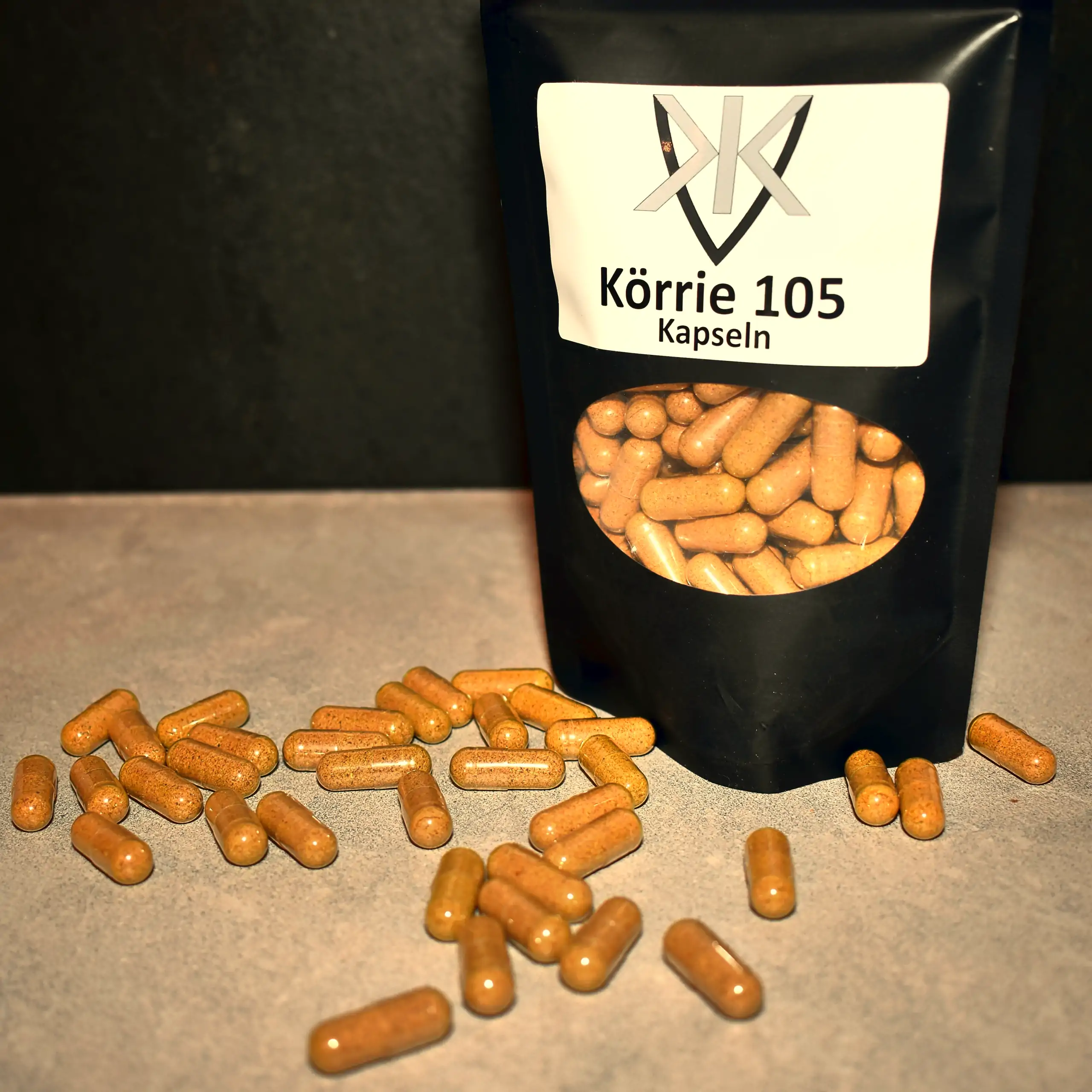 Ergänzende Ansicht Körrie 105 in Kapseln (Körrie 105K): Ein schwarzer Beutel mit transparentem Sichtfenster, gefüllt mit goldenen veganen Kapseln, aus dem einige Kapseln herausfallen und verstreut auf einer grauen, seidenglämzend polierten Marmoroberfläch