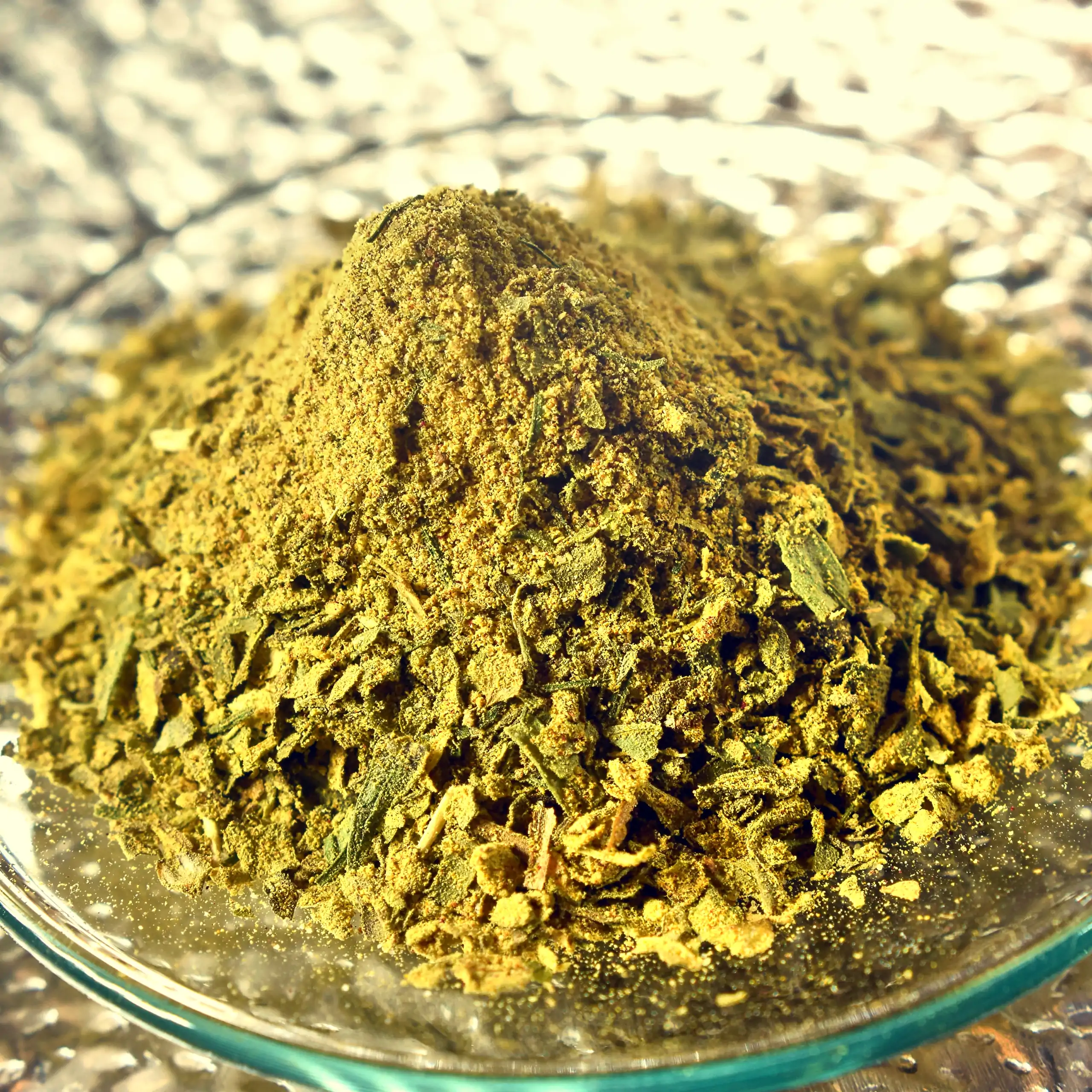 Detailaufnahme der Mischung Körrie 4x4: Ein Haufen leuchtend grüner, grob gemahlener würziger Kräuter- und Gewürzmischung mit feinpulvrigen Anteilen auf einer flachen Glasschale auf silbrig reflektierendem Hintergrund, der die natürliche Textur aus Blätte