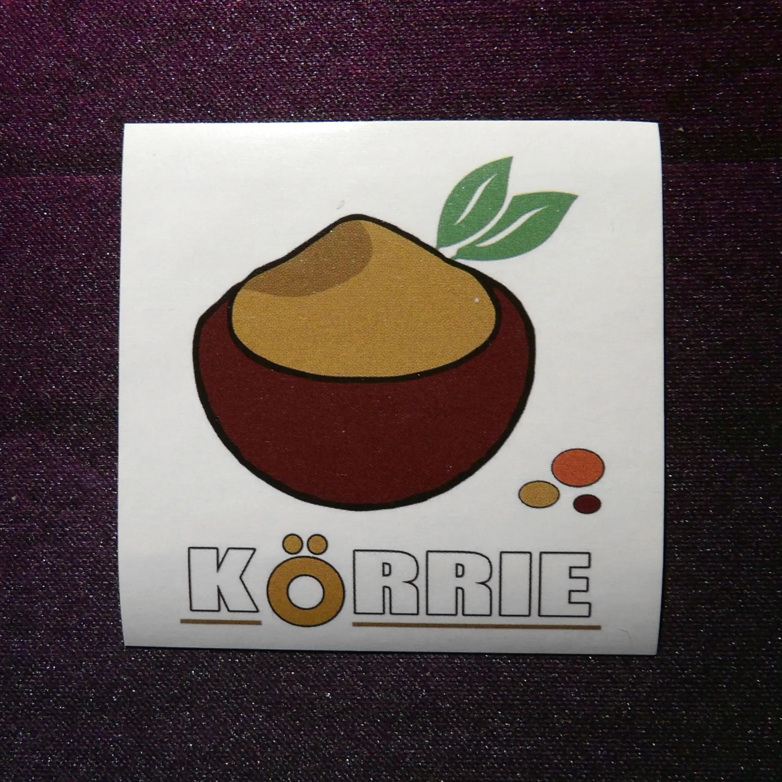 Originalaufkleber (Körrie KFA) "Körrie-Logo" auf dem Trägermaterial: Eine dunkelbraune Schale mit ockerfarbenem Gericht, aus dem zwei grüne Lorbeerblätter mit feinen Konturlinien ragen, symbolisierend Frische und natürliche Reinheit; neben der Schale lieg