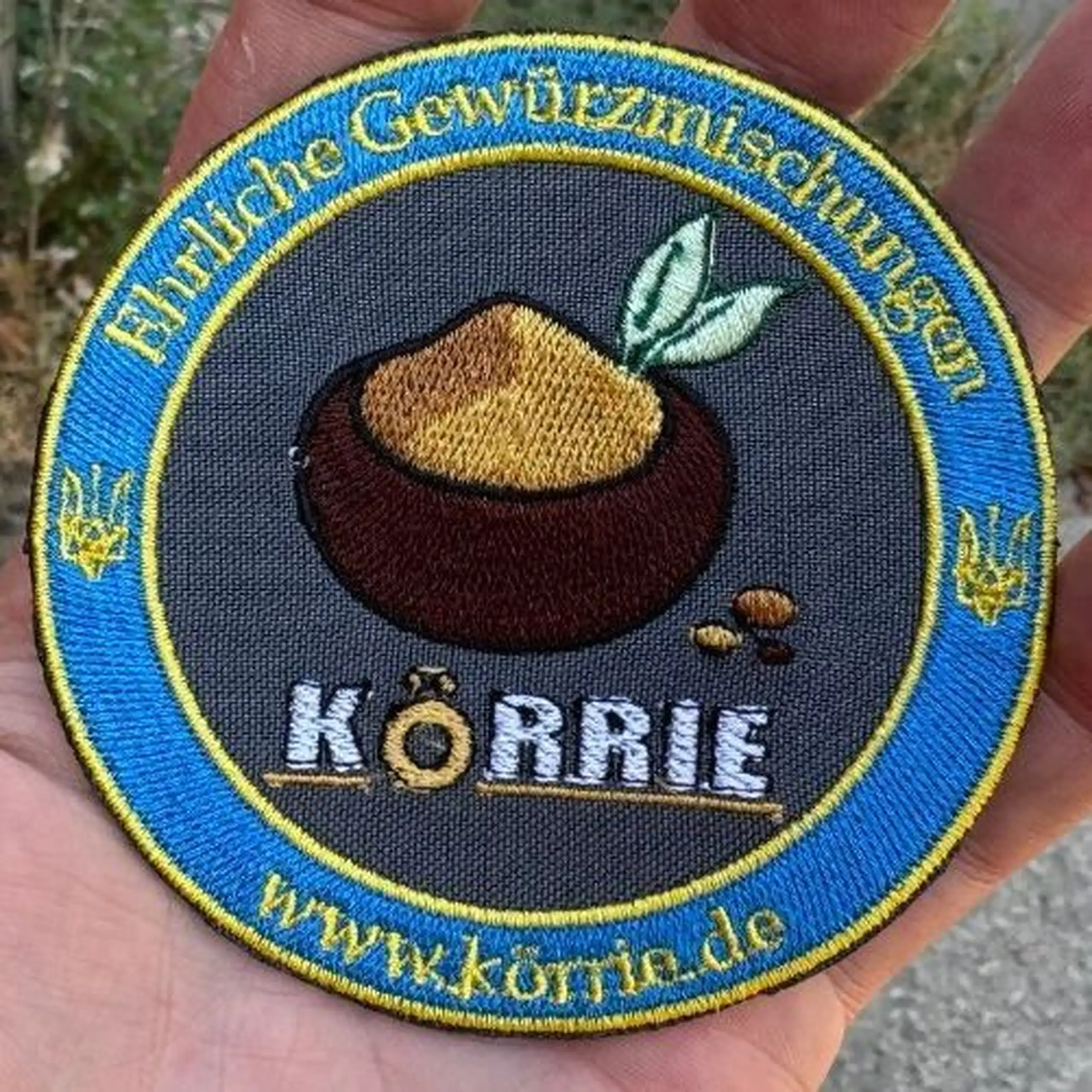 Detailaufnahme des gestickten Körrie-Fan-Patch (KFP), hier in der Version "dark" mit dunkelgrauem Hintergrund. Handgefertigt in der Ukraine, mit dem Logo ehrlicher Gewürzmischungen aus Berlin: braune Schale mit ockerfarbenem Gericht, grünen Lorbeerblätter