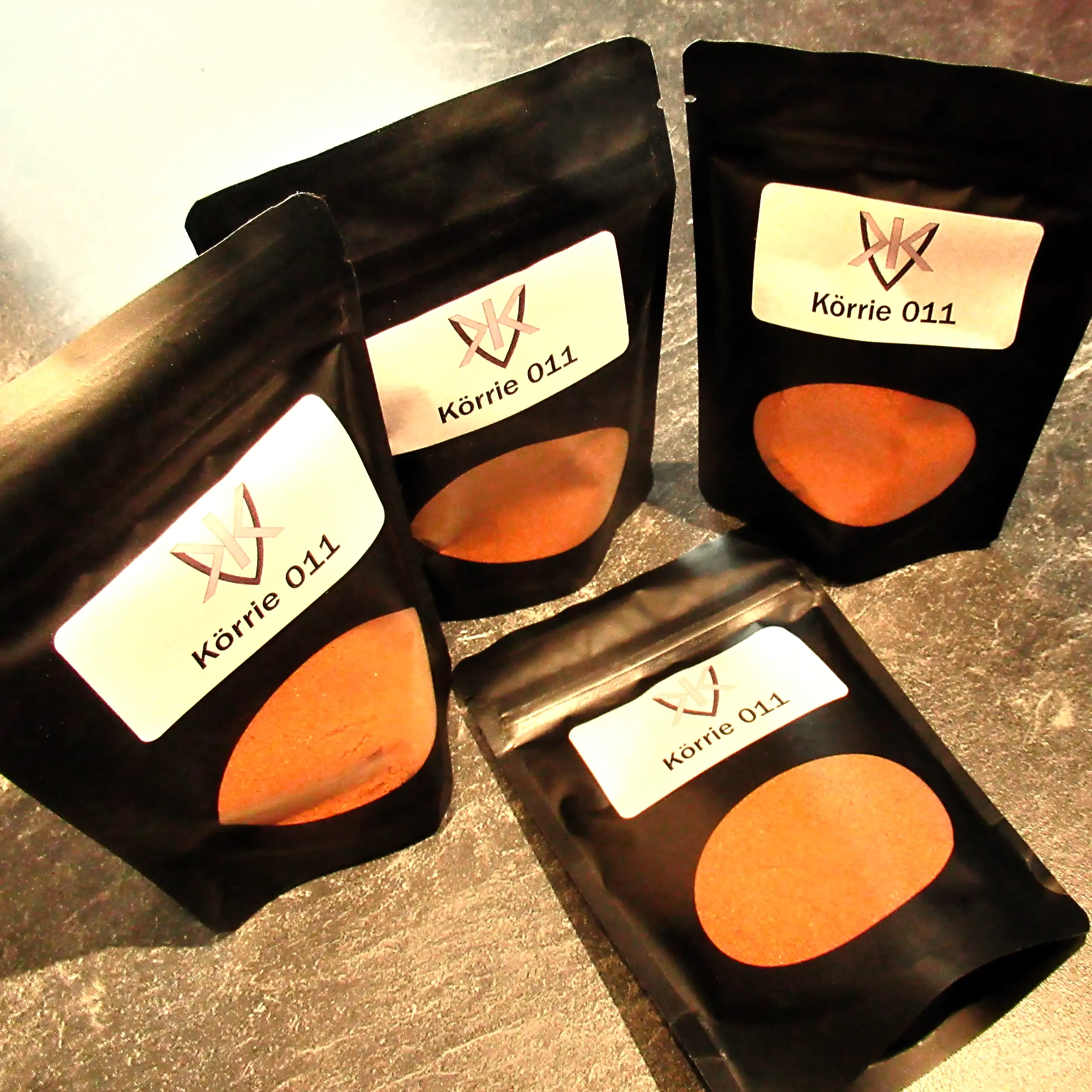 Zusätzliches Bild der Körrie 011 Gewürzmischung: Vier schwarze Beutel mit transparentem Sichtfenster, durch das die leuchtend kräftig-orangene, fein gemahlene rauchig-scharfe Mischung sichtbar ist, versehen mit weißen Labels "Körrie 011" und dem Logo von 