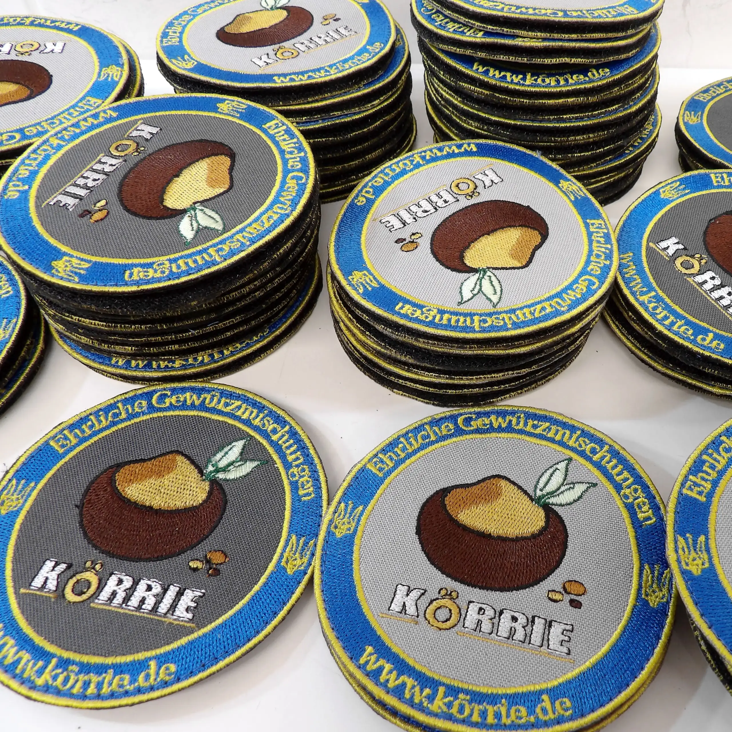 Ergänzende Ansicht der gestickten Körrie-Fan-Patches "Zeig Deine Liebe zu Körrie!", handgefertigt in der Ukraine, mit dem Logo ehrlicher Gewürzmischungen aus Berlin: braune Schale mit ockerfarbenem Gericht, grünen Lorbeerblättern und bunten Gewürzkörnern,