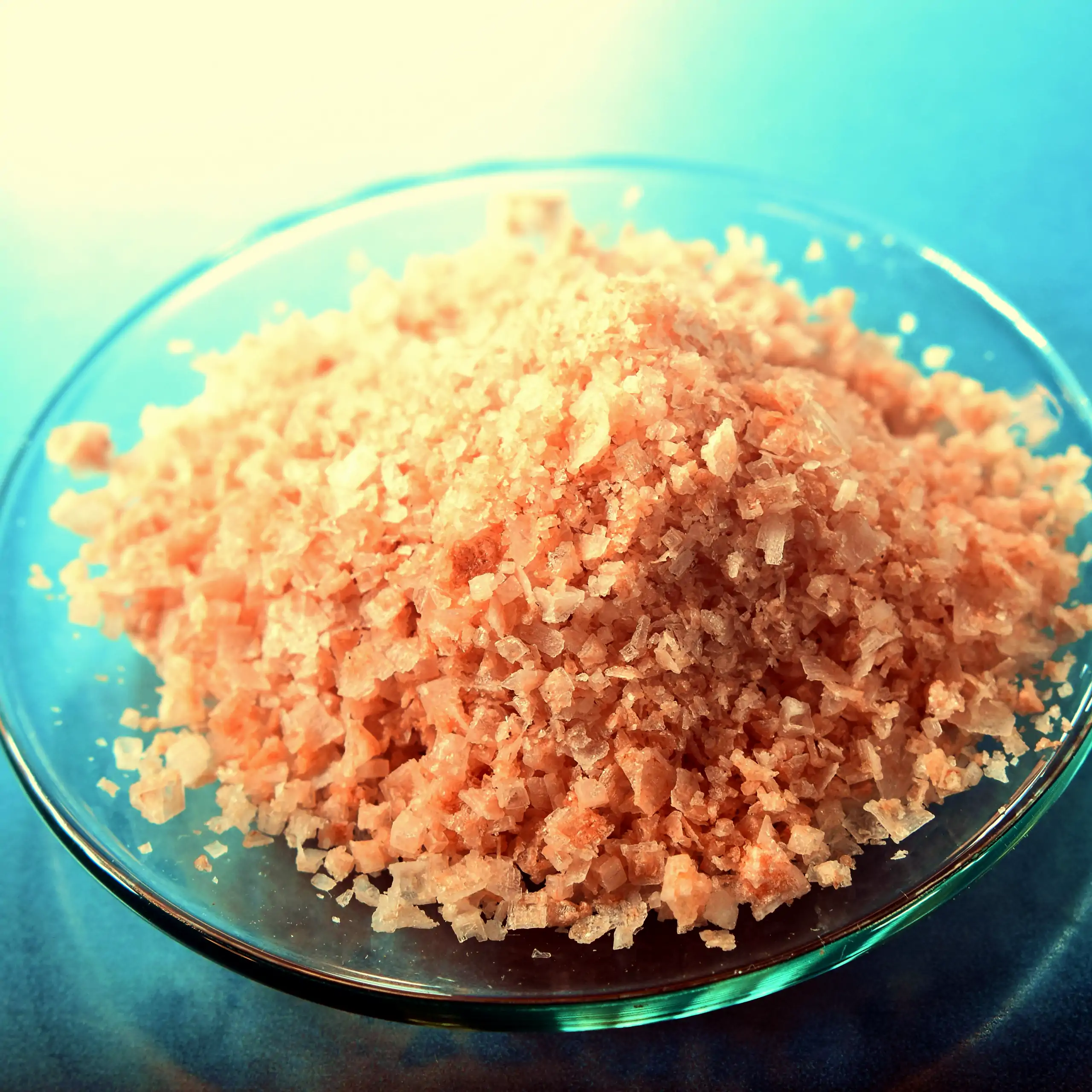 Detailaufnahme der rosa Salzflocken: Ein Haufen leuchtend rosa, knusprig-flockiger Salzmischung in einer transparenten Glasschale auf blauem Hintergrund, der die mineralreiche, kristalline Textur und natürliche Färbung betont für einen frischen, appetitli