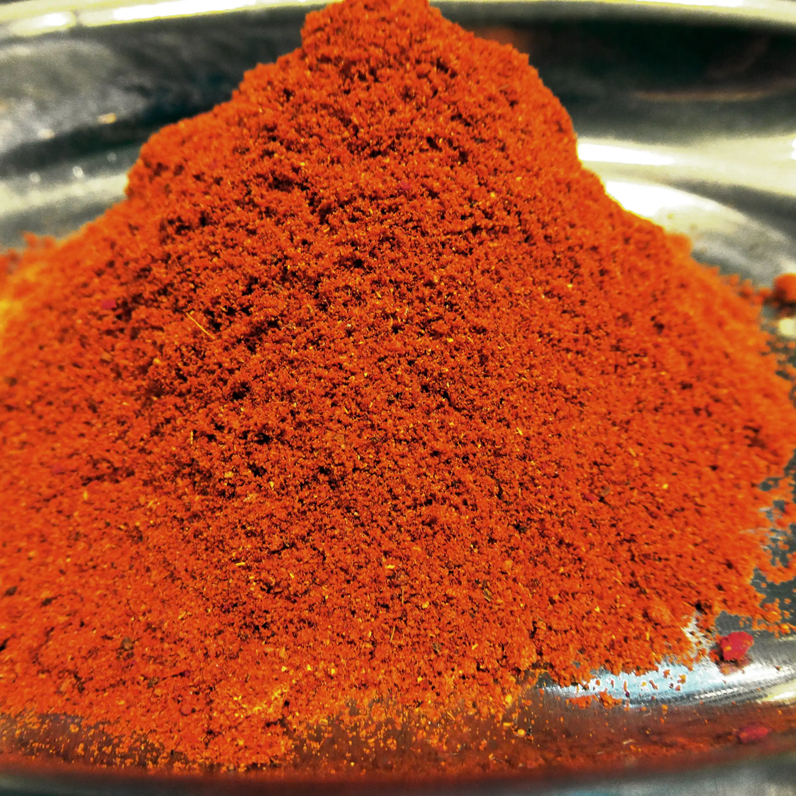 Ergänzende Ansicht der Körrie U80 Gewürzmischung: Ein Haufen leuchtend orangener, fein gemahlener scharfer Gewürzmischung in einer metallischen Schale auf dunklem Hintergrund. Nahaufnahme, die intensive, feurige Textur und rauchige Nuance betonend für ein