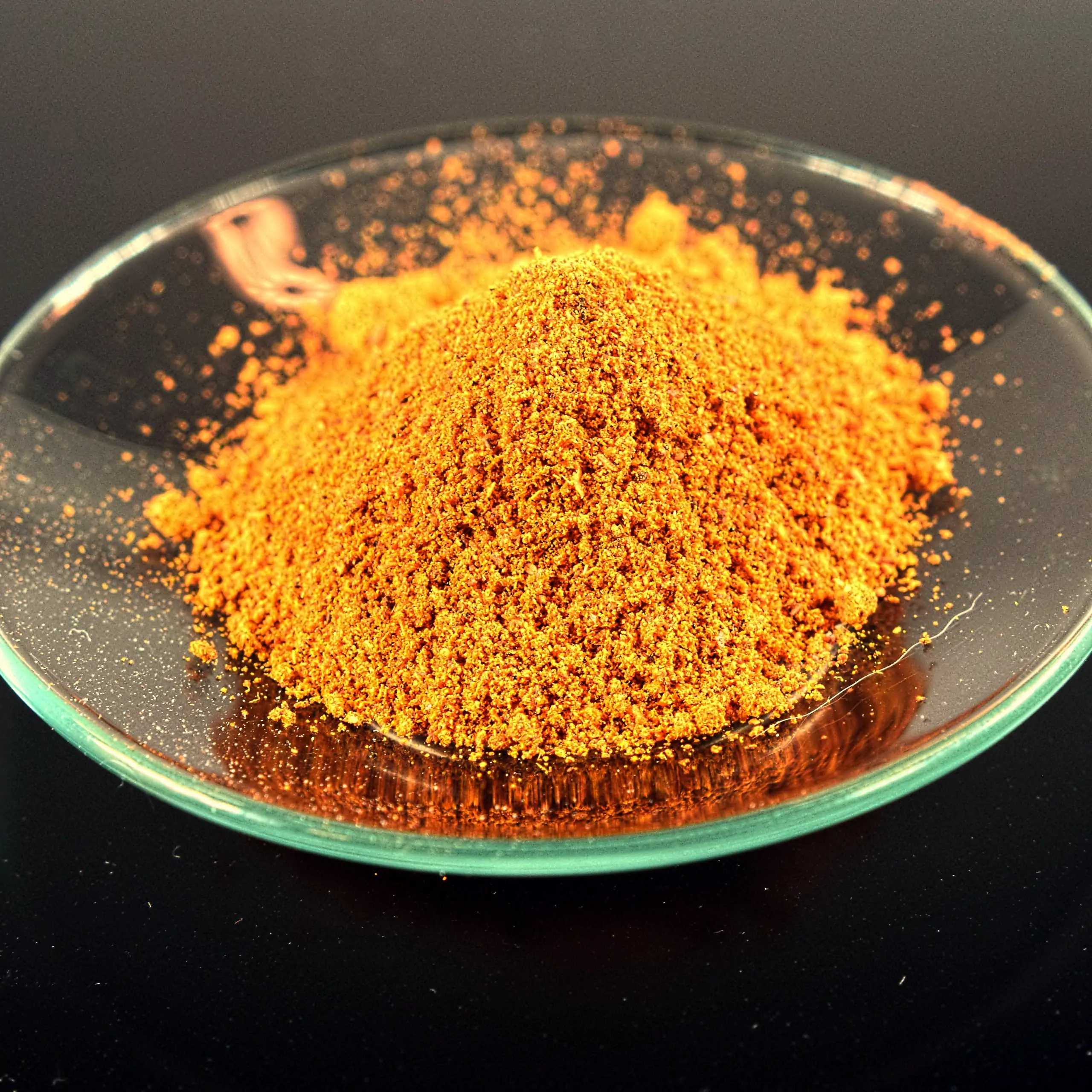 Detailaufnahme der Körrie H55 Gewürzmischung: Ein sorgfältig drapierter Haufen leuchtend orangener, fein gemahlener, herzhafter Gewürzmischung in einer transparenten Glasschale auf dunklem Hintergrund, der die intensive, aromatische Textur und vanillige N