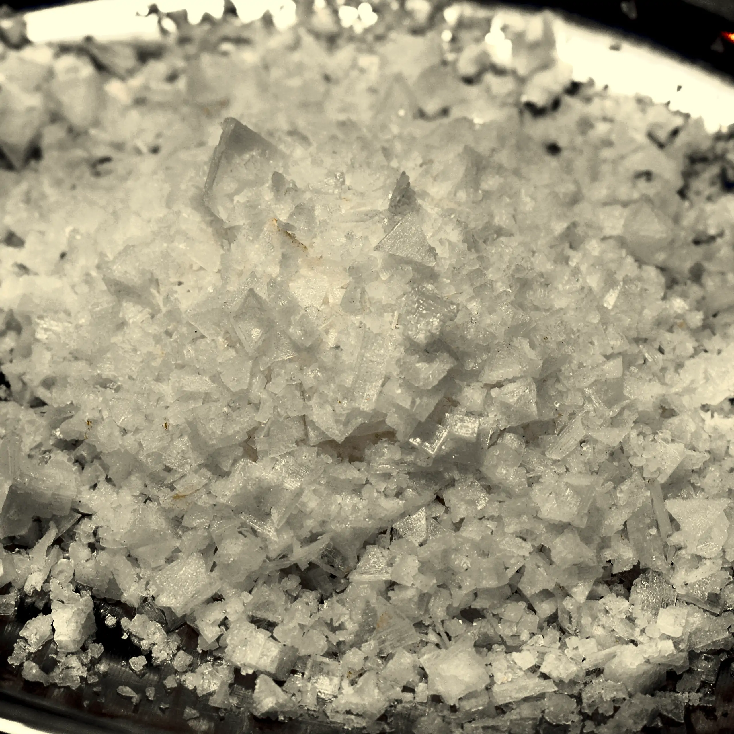 Ergänzende Ansicht der Fleur de Sel (Pyramidensalz): Ein Haufen leuchtend weißer, knuspriger Salzflocken mit pyramidenförmiger Kristallstruktur in einer Edelstahl vor dunklem Hintergrund, der die kristalline Struktur und natürliche Reinheit dieses Meersal