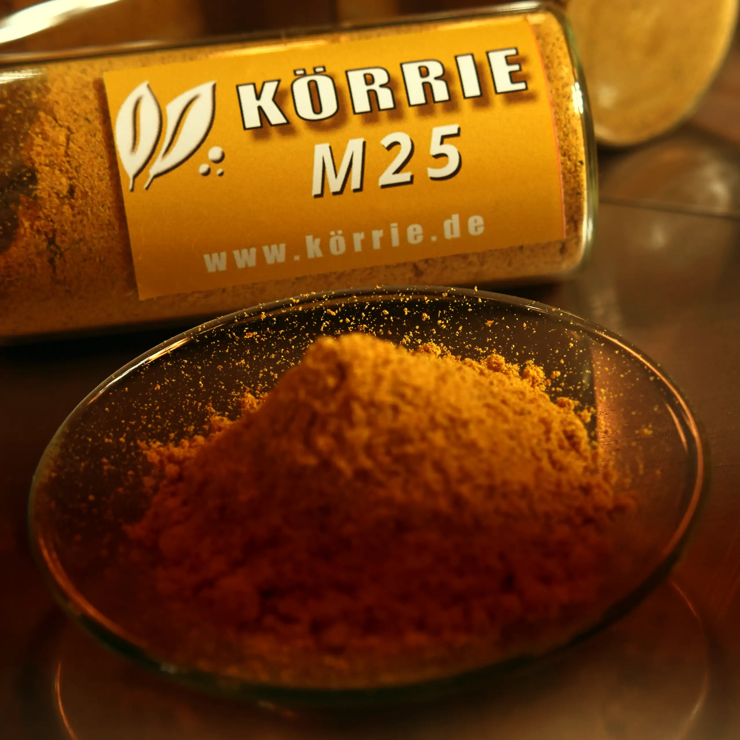 Zusätzliches Bild der Körrie M25 Gewürzmischung: Eine leuchtend goldgelbe, fein gemahlene vollmundige Würzmischung in einem transparenten Glasgefäß auf der Seite liegend, mit gelbem Label "KÖRRIE M25" und Website www.körrie.de, neben einer Glasschale mit 