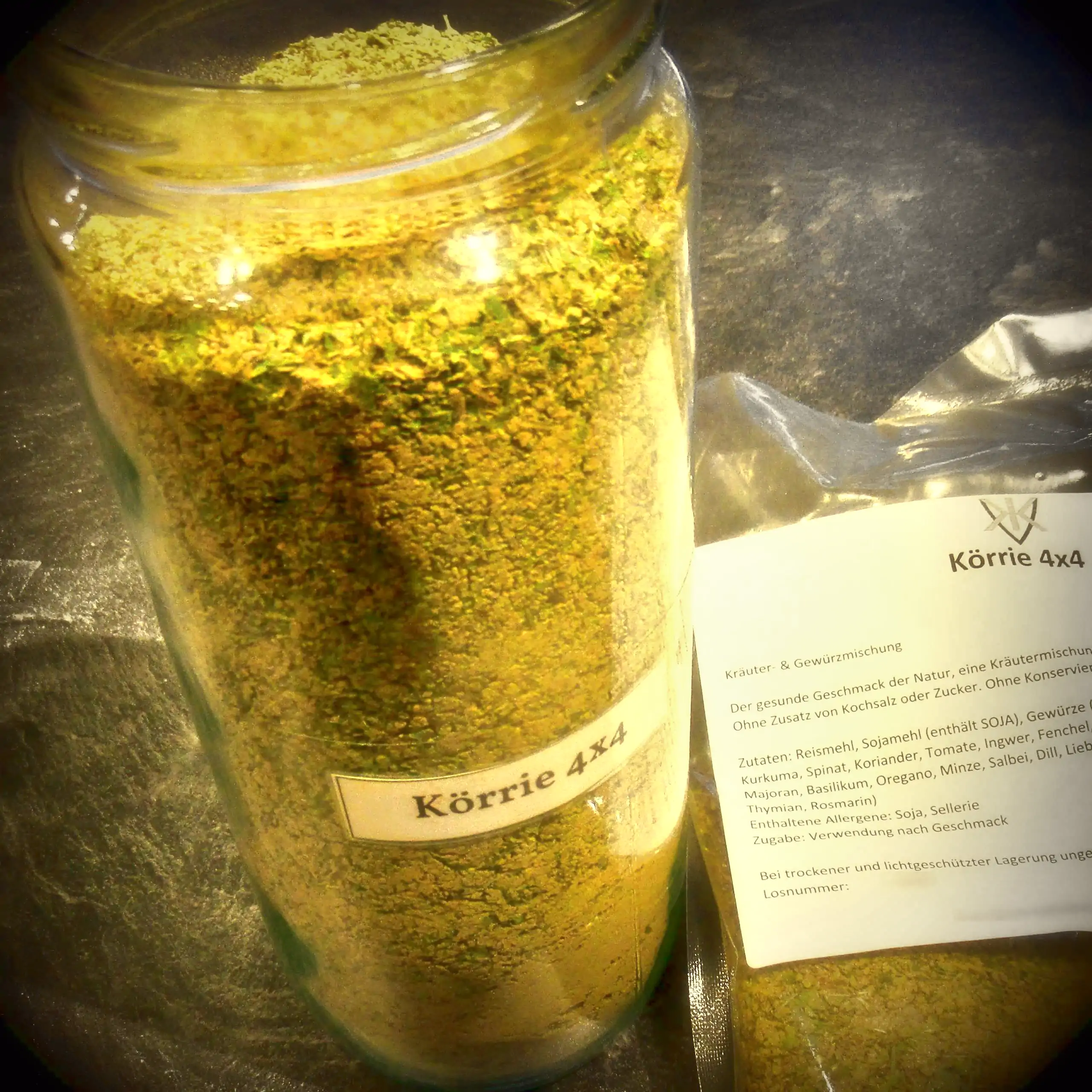 Alternatives Vorschaubild der Galerie für die Kräuter- und Gewürzmischung Körrie 4x4: Ein hohes, transparentes Glasgefäß gefüllt mit leuchtend grüner, grob gemahlener Mischung mit feinen Anteilen, versehen mit einem weißen Label "Körrie 4x4", neben einem 