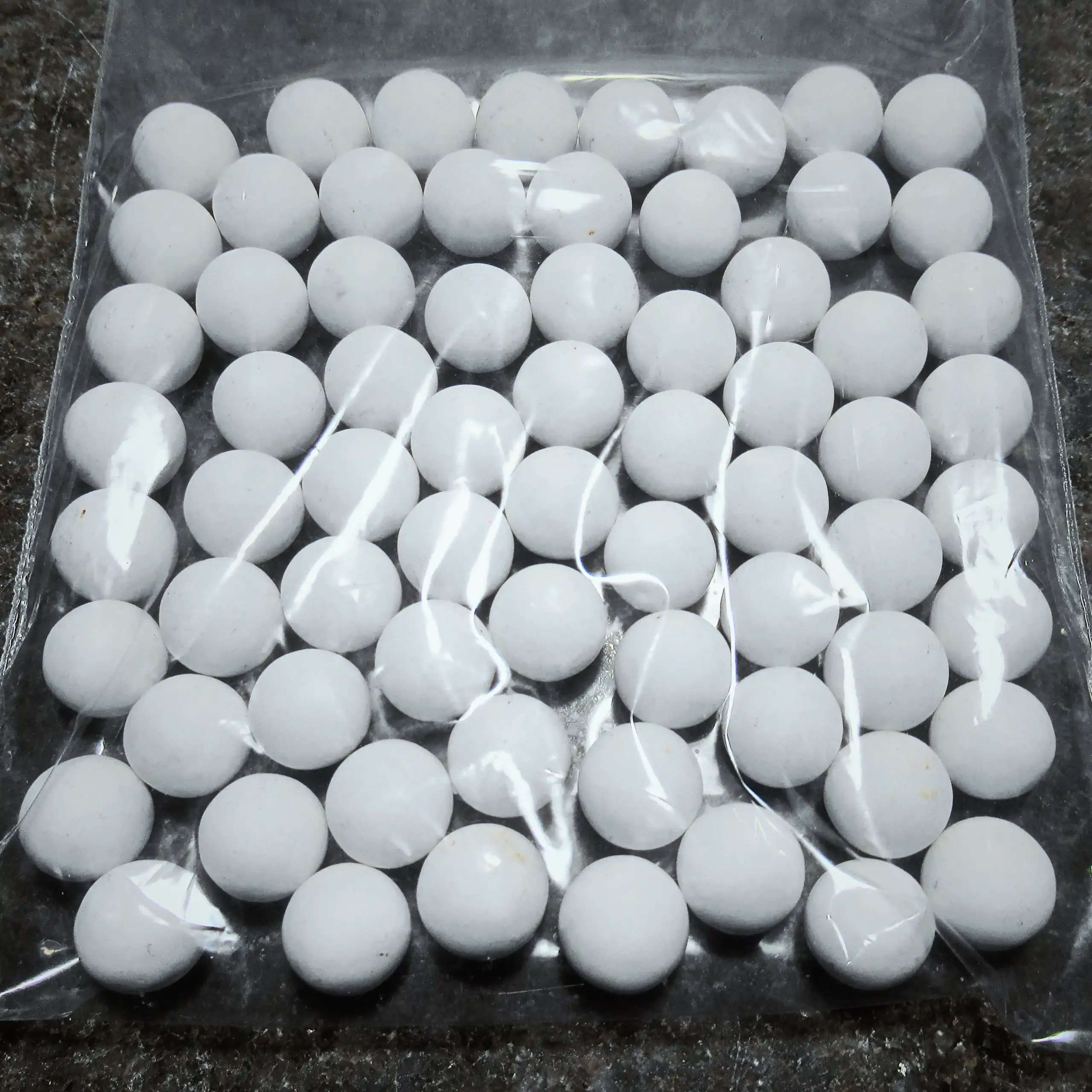 Zusätzliches Bild der Körrie-Aktiv-Perlen (Keramik) / Körrie KAP S: Eine große Menge leuchtend weißer, mattierter, runder Keramikperlen aus Aluminiumoxid in einer durchsichtigen Plastikverpackung, verstreut auf einer dunklen Oberfläche für eine reine, fun