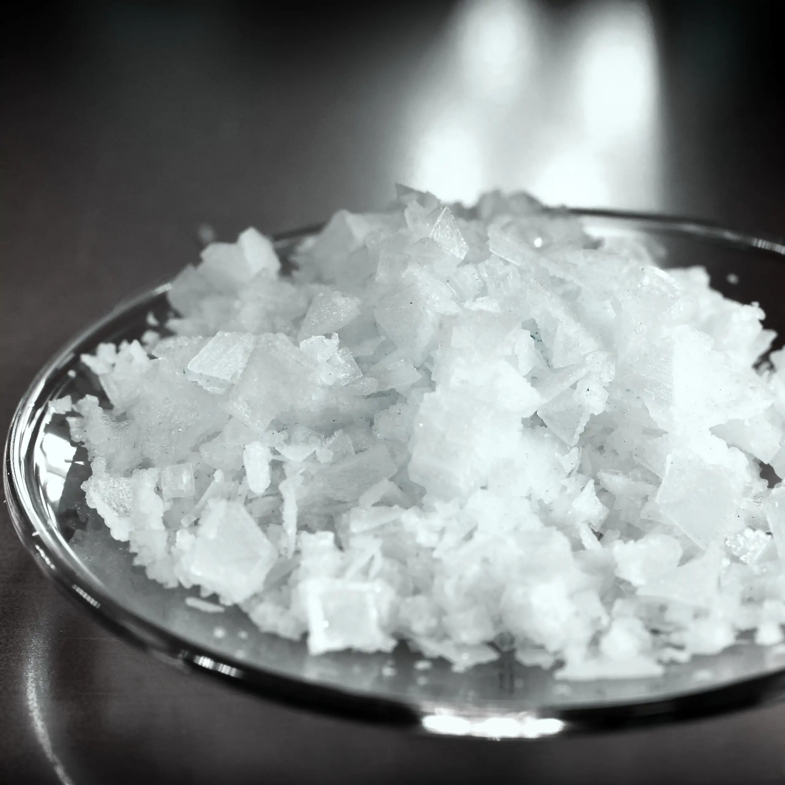 Detailaufnahme der Fleur de Sel Pyramidensalz: Ein Häuflein leuchtend weißer, knusprig-pyramidenförmiger, fein-knackiger Salzkristalle in einer transparenten Glasschale auf dunklem Hintergrund, der die mineralreiche, kristalline Struktur und natürliche Re