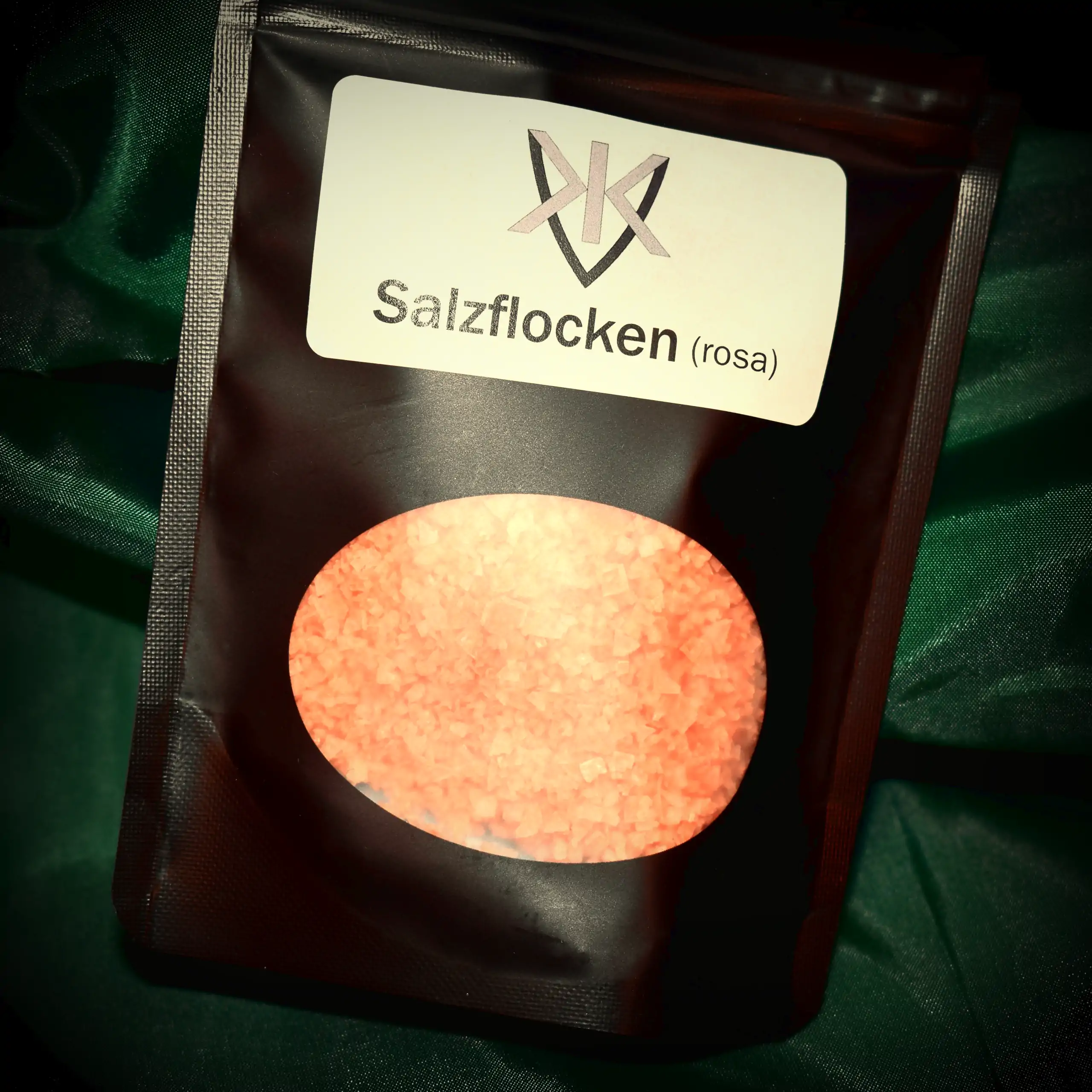 Alternatives Vorschaubild der Galerie für rosa Salzflocken: Ein schwarzer Beutel mit transparentem Sichtfenster, durch das die leuchtend rosa-farbenen, knusprig-flockigen Salzkristalle sichtbar sind, versehen mit einem weißen Label, das das KVK-Logo und "