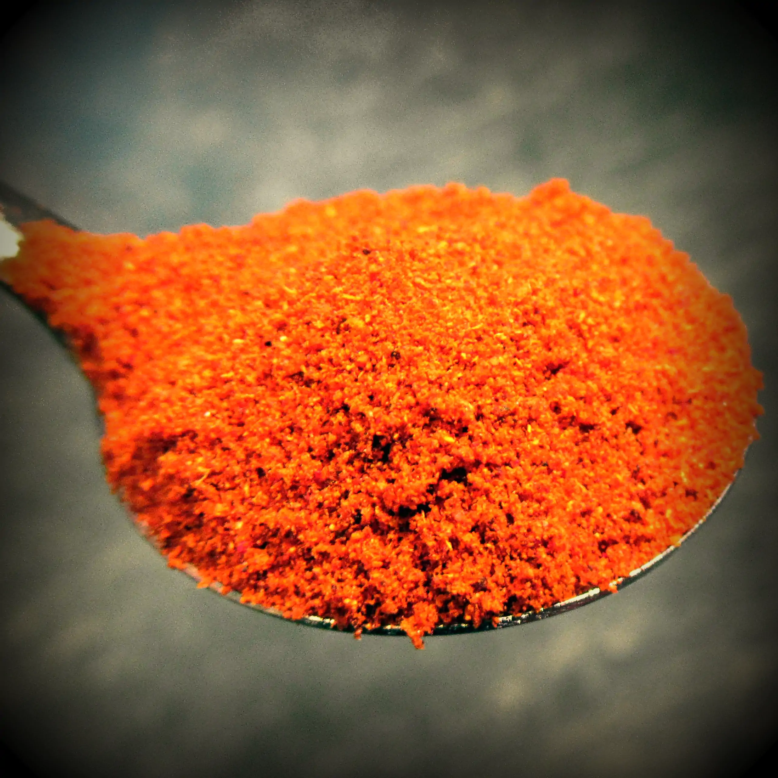 Zusätzliches Bild der Körrie U80 Gewürzmischung: Ein Haufen leuchtend orangener, fein gemahlener scharfer Gewürzmischung auf einem Löffel auf dunklem Hintergrund mit Vignetten-Effekt, der die intensive, feurige Textur und rauchige Nuance betont für einen 