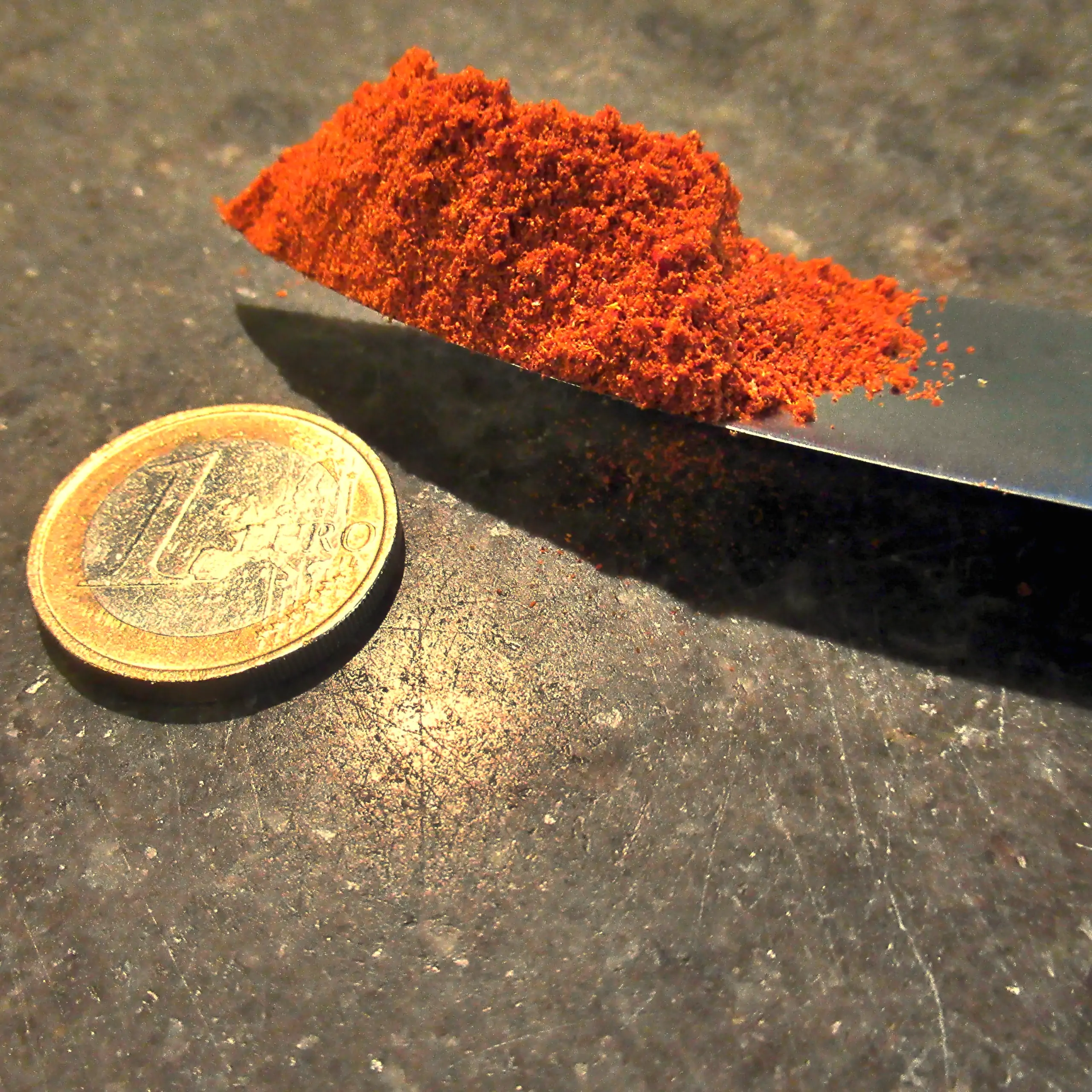 Alternatives Vorschaubild der Galerie für Körrie 8zB Gewürzmischung: Ein Haufen kräftig leuchtend-roter, fein gemahlener, aromatischer Gewürzmischung auf der Klinge eines Messers, neben einer 1-Euro-Münze zum Maßstabsvergleich, präsentiert auf einer dunkl