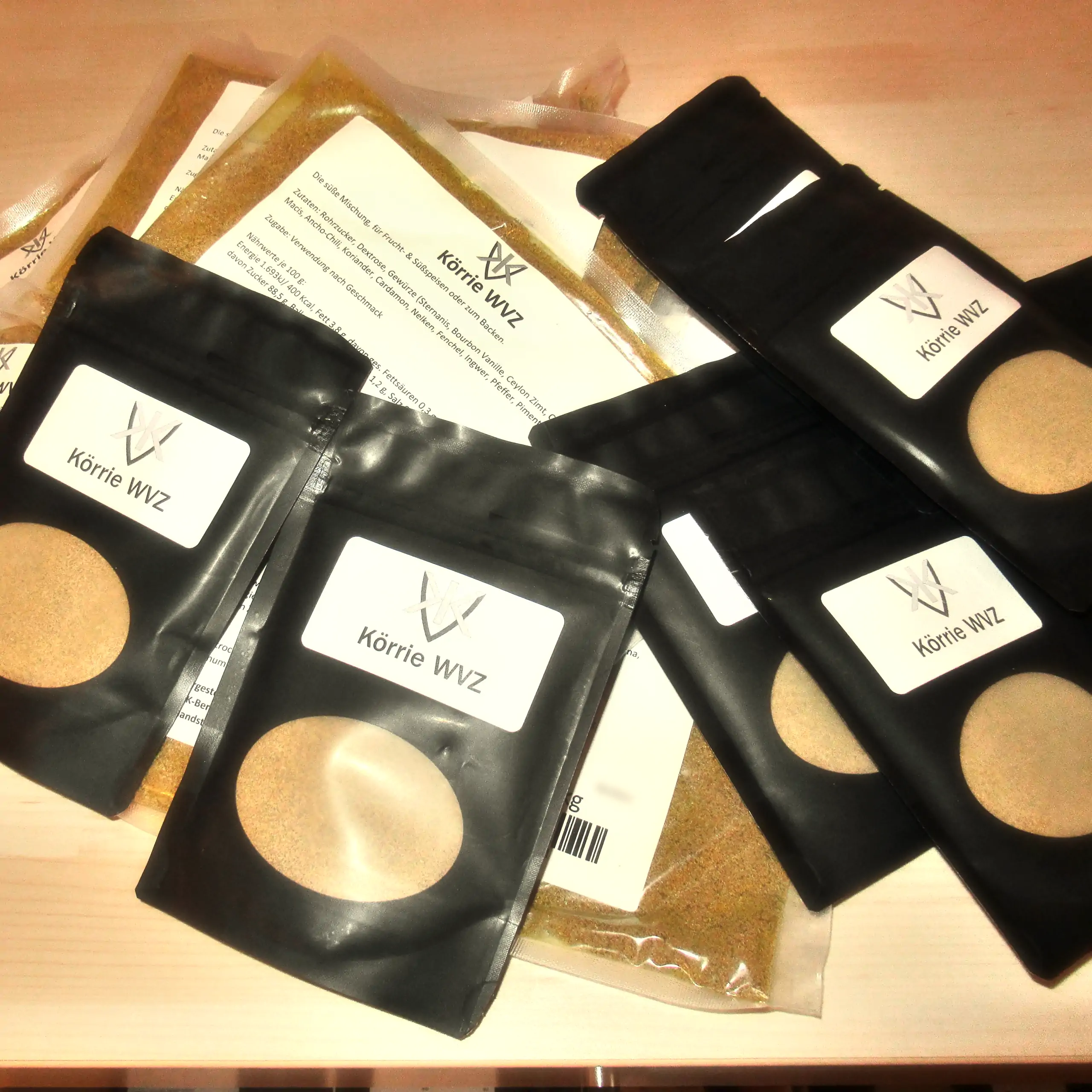 Ergänzende Ansicht der Mischung Körrie WVZ: Mehrere schwarze Beutel mit transparentem Sichtfenster, durch das die leuchtend goldbraune, fein gemahlene würzig-süße Mischung Körrie WVZ mit sichtbaren Rohrzuckerkristallen erkennbar ist, versehen mit weißen L