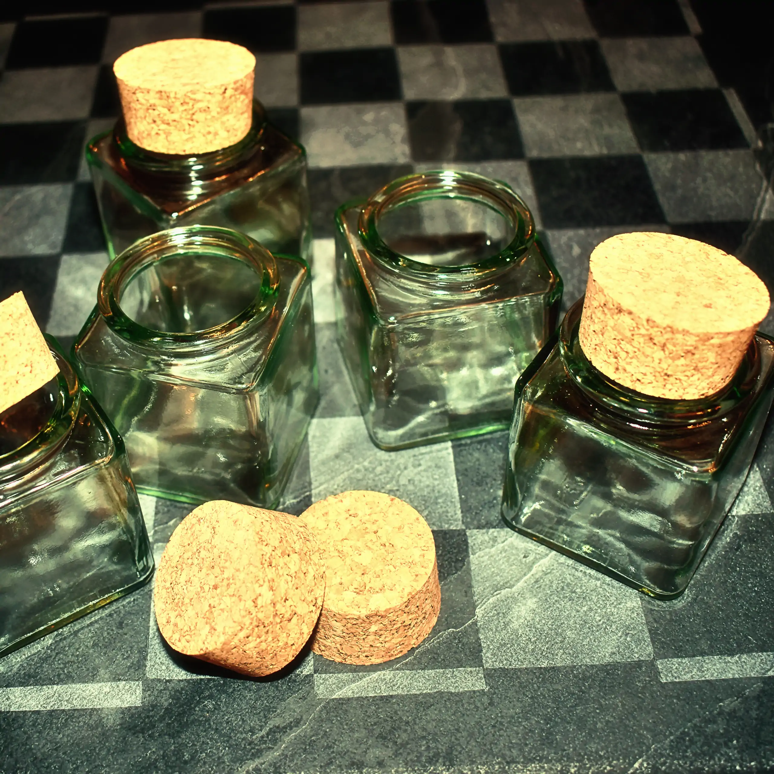 Ergänzende Ansicht der Körrie-Korken-Gläser: Eine Gruppe von eleganten, quadratischen Glasgefäßen mit natürlichen Korkverschlüssen, einige offen, einige geschlossen, und lose Korken davor, präsentiert auf einer schachbrettartigen Oberfläche aus graviertem