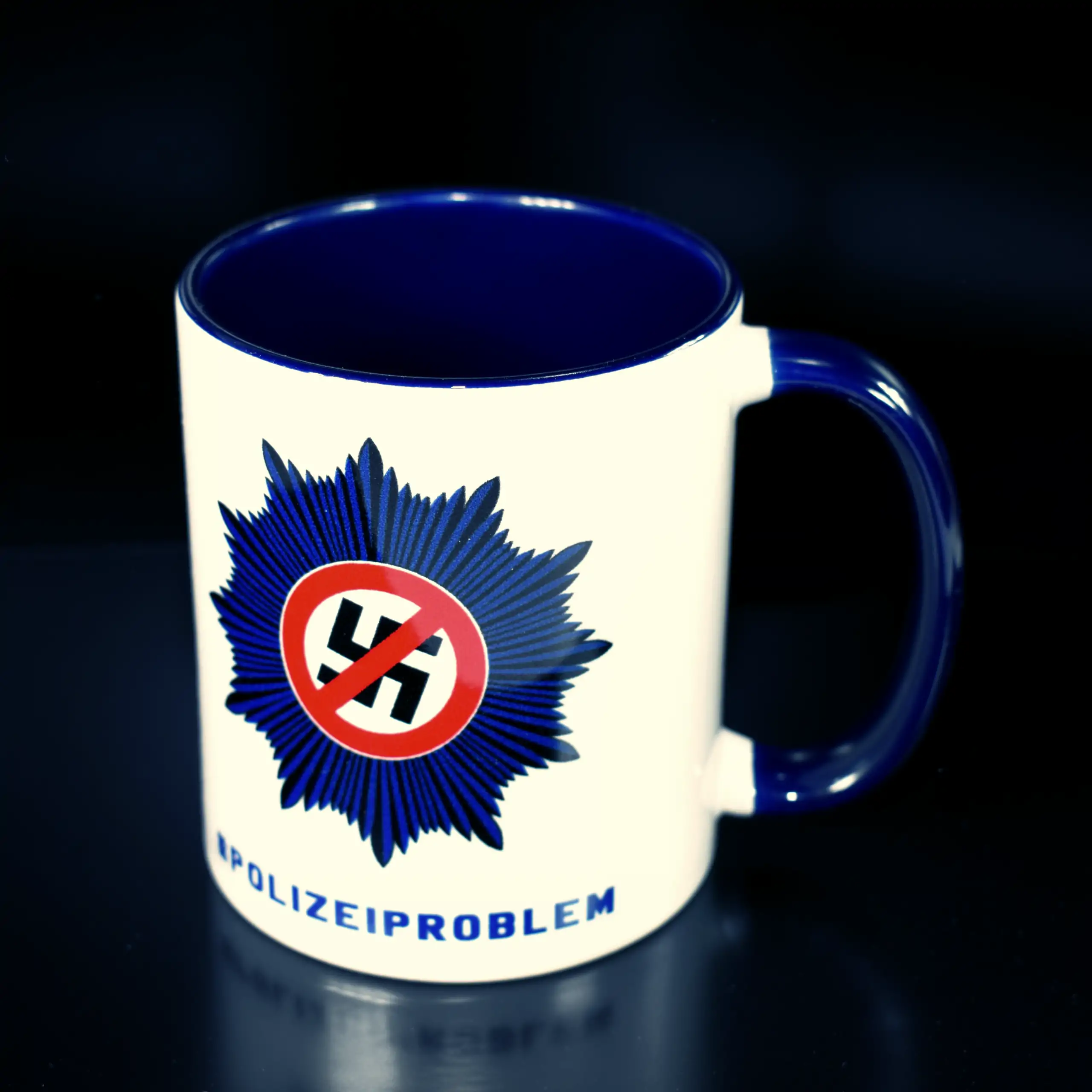 Titelbild der Tasse "Nazis raus aus der Polizei" (Variante: Griff rechts): Eine weiße Keramiktasse mit blauem Innenrand und Henkel, präsentiert auf einem dunklen Untergrund für einen kontrastreichen, markanten Look, wobei der Henkel rechts vom Logo positi