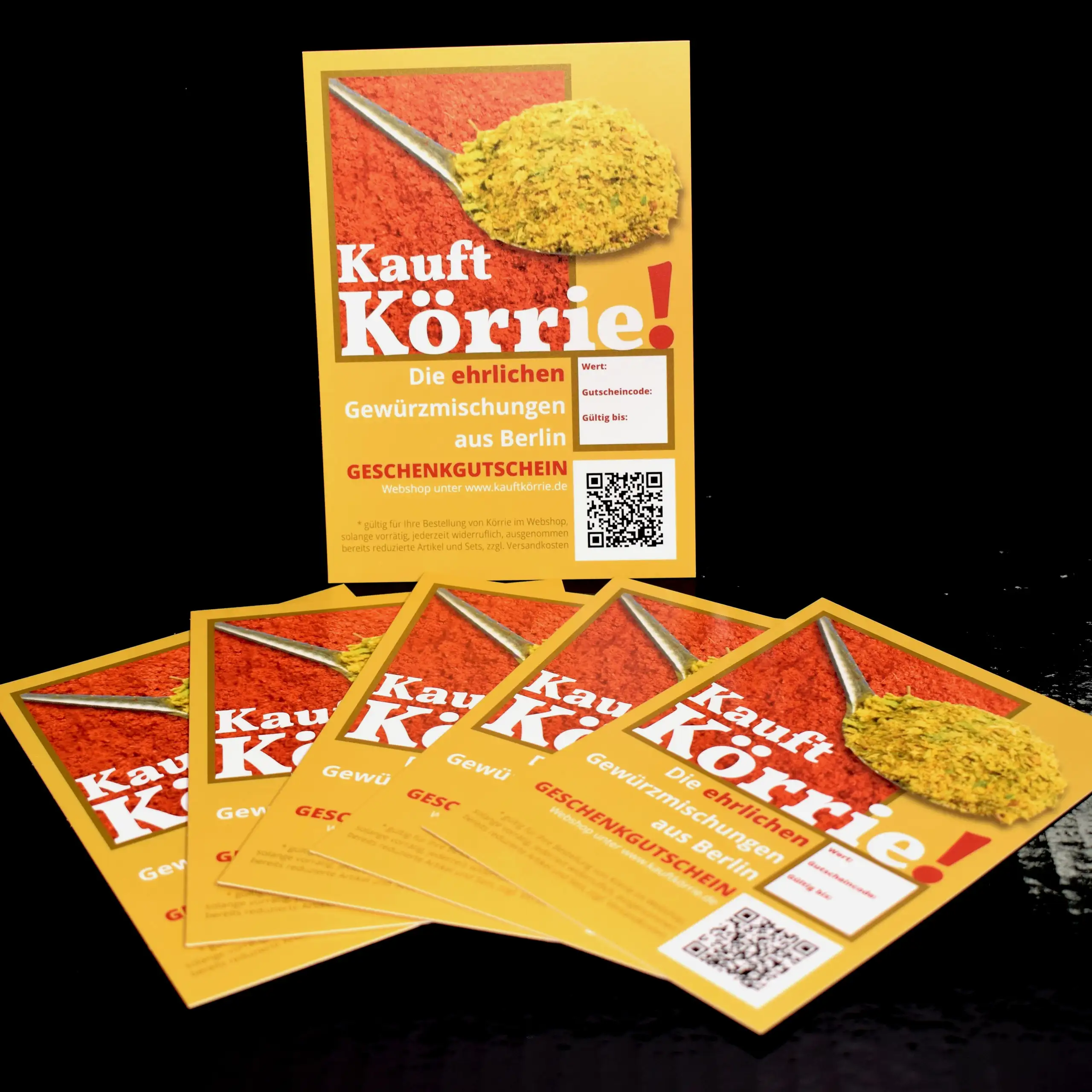 Alternatives Vorschaubild der Galerie für Körrie-Geschenkgutschein / Körrie KGG: Ein Stapel leuchtend gelber Geschenkgutscheine, fächerförmig arrangiert auf einem dunklen Hintergrund - im Hintergrund ein aufrecht gestellter Gutschein mit dem prominenten C