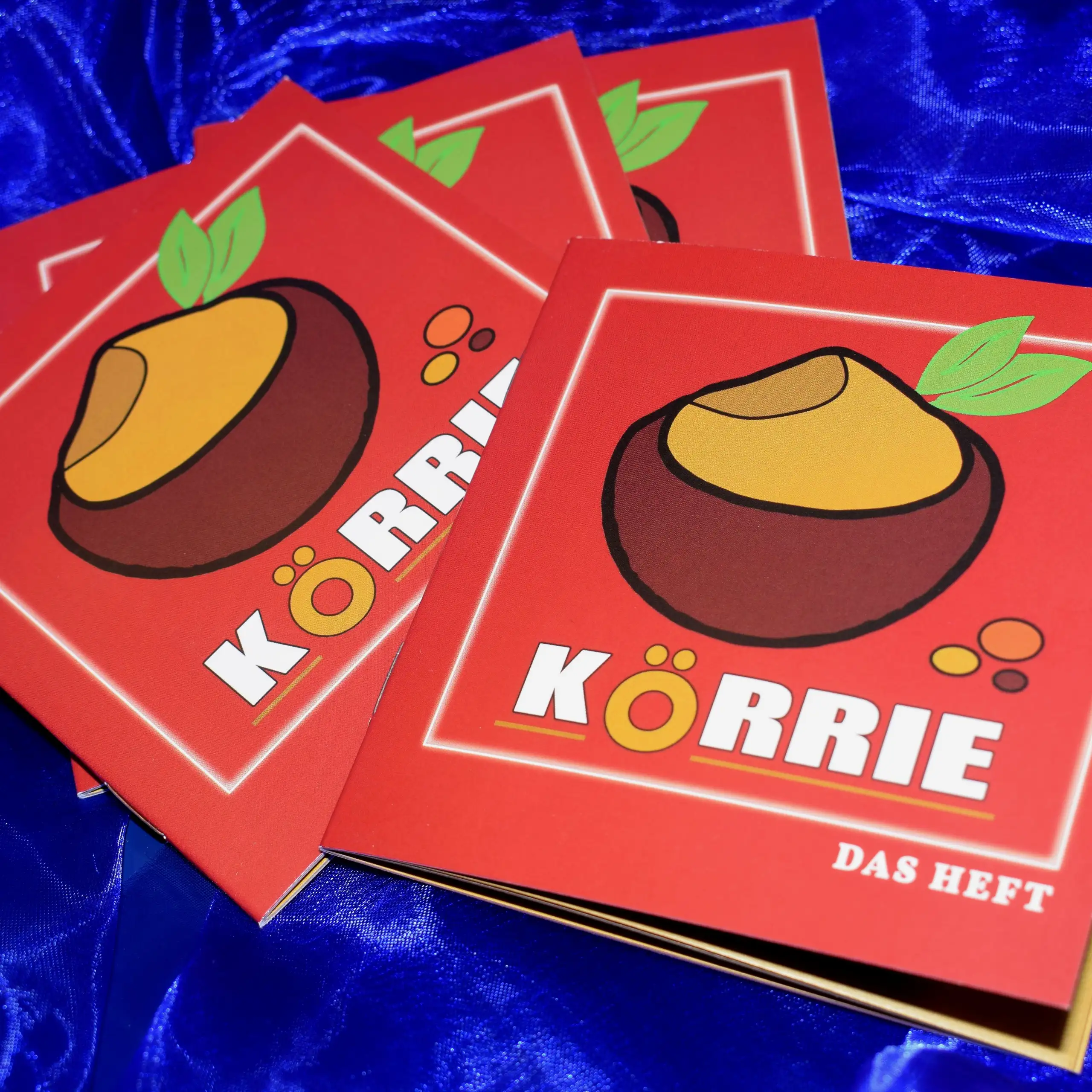 Alternatives Vorschaubild der Galerie für Körrie-Support-Heft / Körrie KSH: Ein Stapel roter Hefte mit dem Markenlogo von Körrie, einer Schale mit einem beigefarbenem Gericht, aus dem grüne Blätter ragen Blättern, dem Titel "KÖRRIE - DAS HEFT" in weißer S