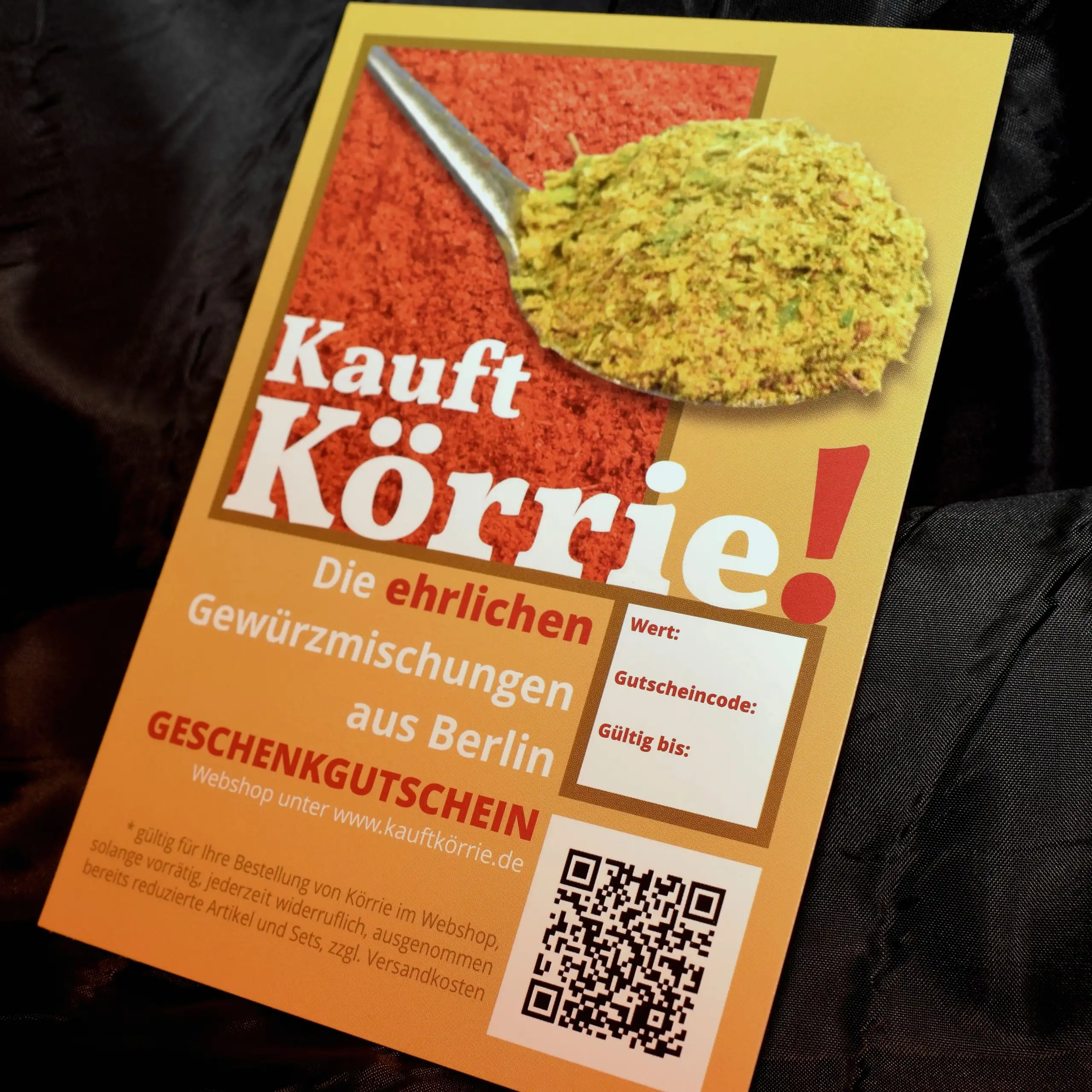 Ergänzende Ansicht des Körrie-Geschenkgutscheins / Körrie KGG: Ein ansprechendes, goldenes Design mit einem prominent in Bild ragenden Löffel voller appetitlicher Gewürzmischung auf rotem Hintergrund, dem prominenten Claim "Kauft Körrie!" in großer weißer