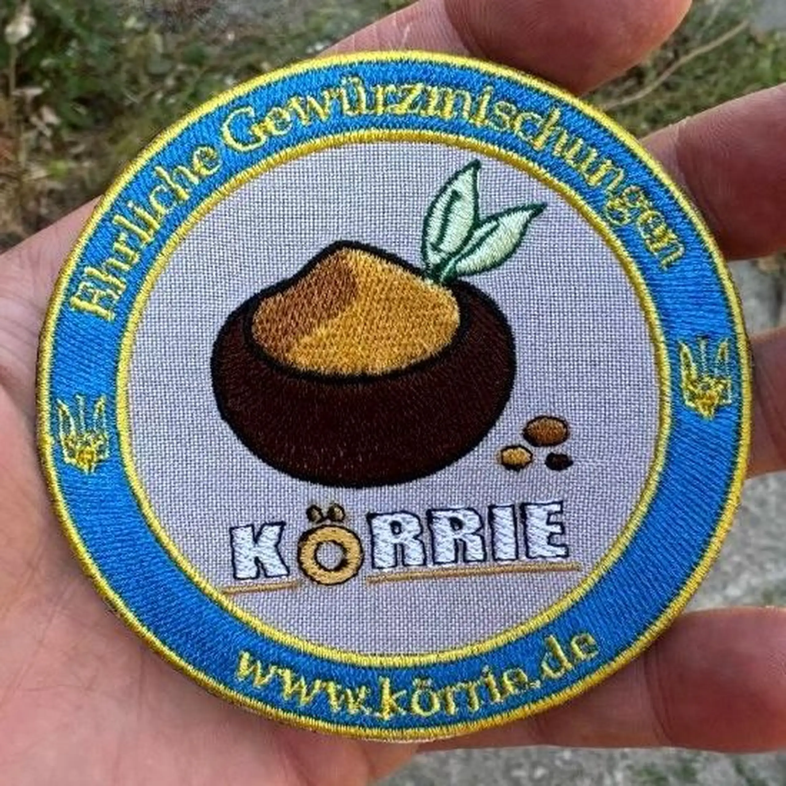 Detailaufnahme des gestickten Körrie-Fan-Patch (KFP), hier in der Version "light" mit hellgrauem Hintergrund. Handgefertigt in der Ukraine, mit dem Logo ehrlicher Gewürzmischungen aus Berlin: braune Schale mit ockerfarbenem Gericht, grünen Lorbeerblättern
