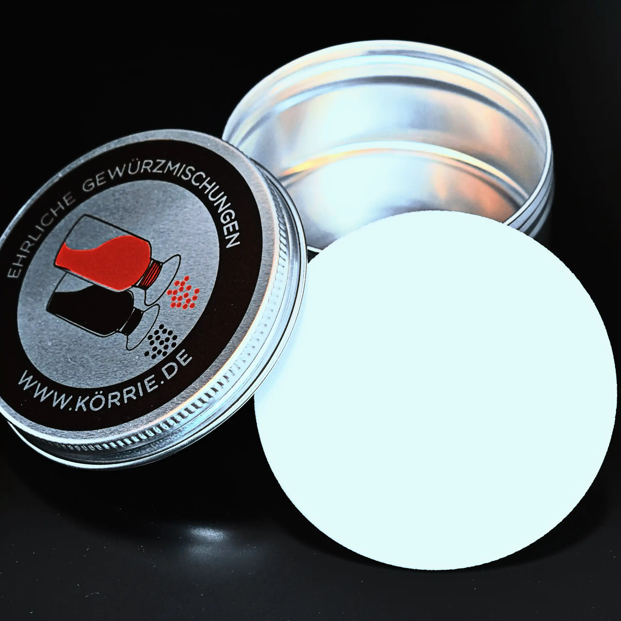 Detailaufnahme der Körrie-Support-Dose: Eine kompakte, runde Metall-Dose aus Aluminium mit Deckel, offen präsentiert mit dem Deckel und Dichtungseinleger daneben, auf schwarzem, seidenmatt glänzenden Hintergrund, auf dem Deckel aufgedruckt das Körrie-Logo