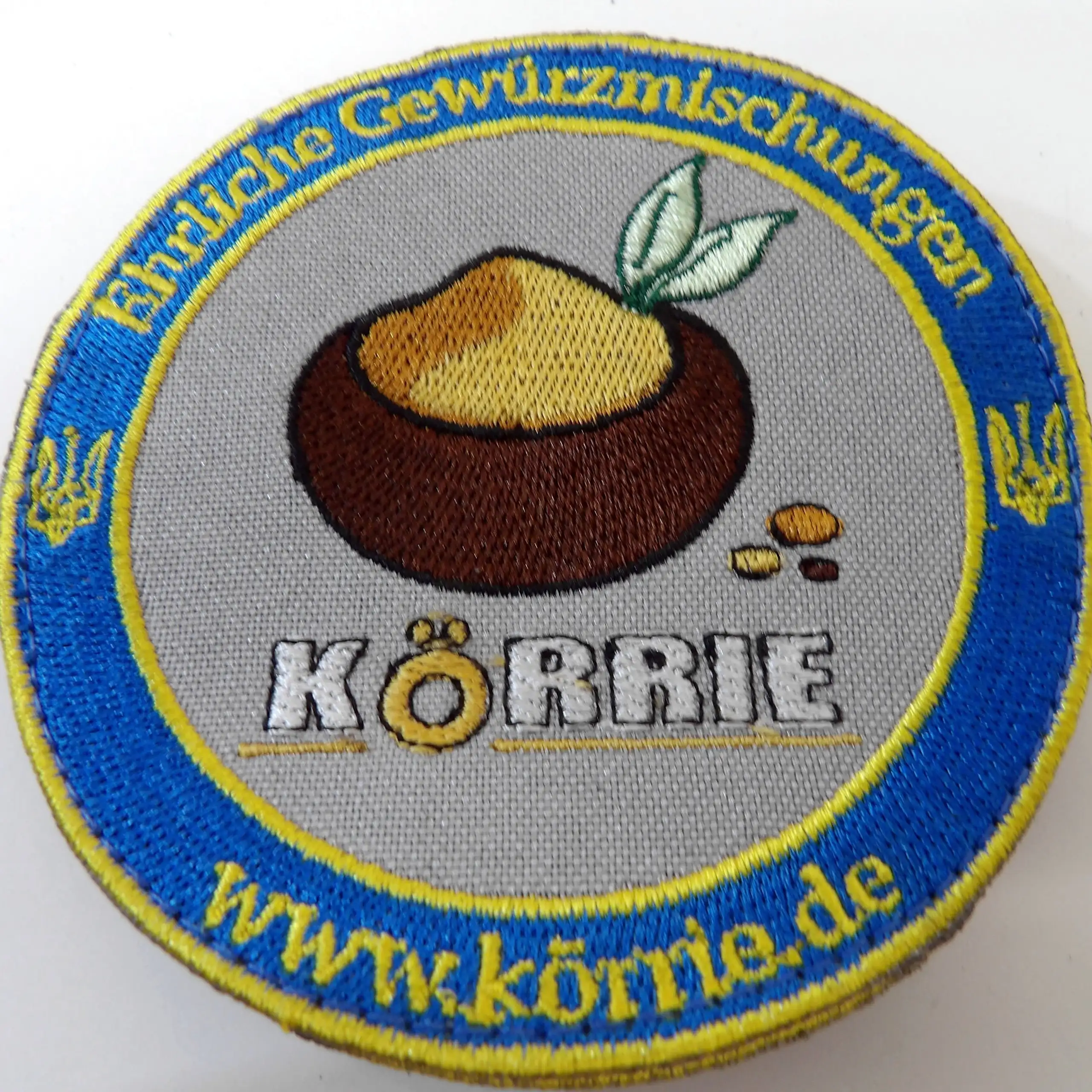 Gestickter Körrie-Fan-Patch (KFP), handgefertigt in der Ukraine, mit dem Logo ehrlicher Gewürzmischungen aus Berlin: braune Schale mit ockerfarbenem Gericht, grünen Lorbeerblättern und bunten Gewürzkörnern, umrahmt vom Schriftzug "KÖRRIE" in weiß mit gold