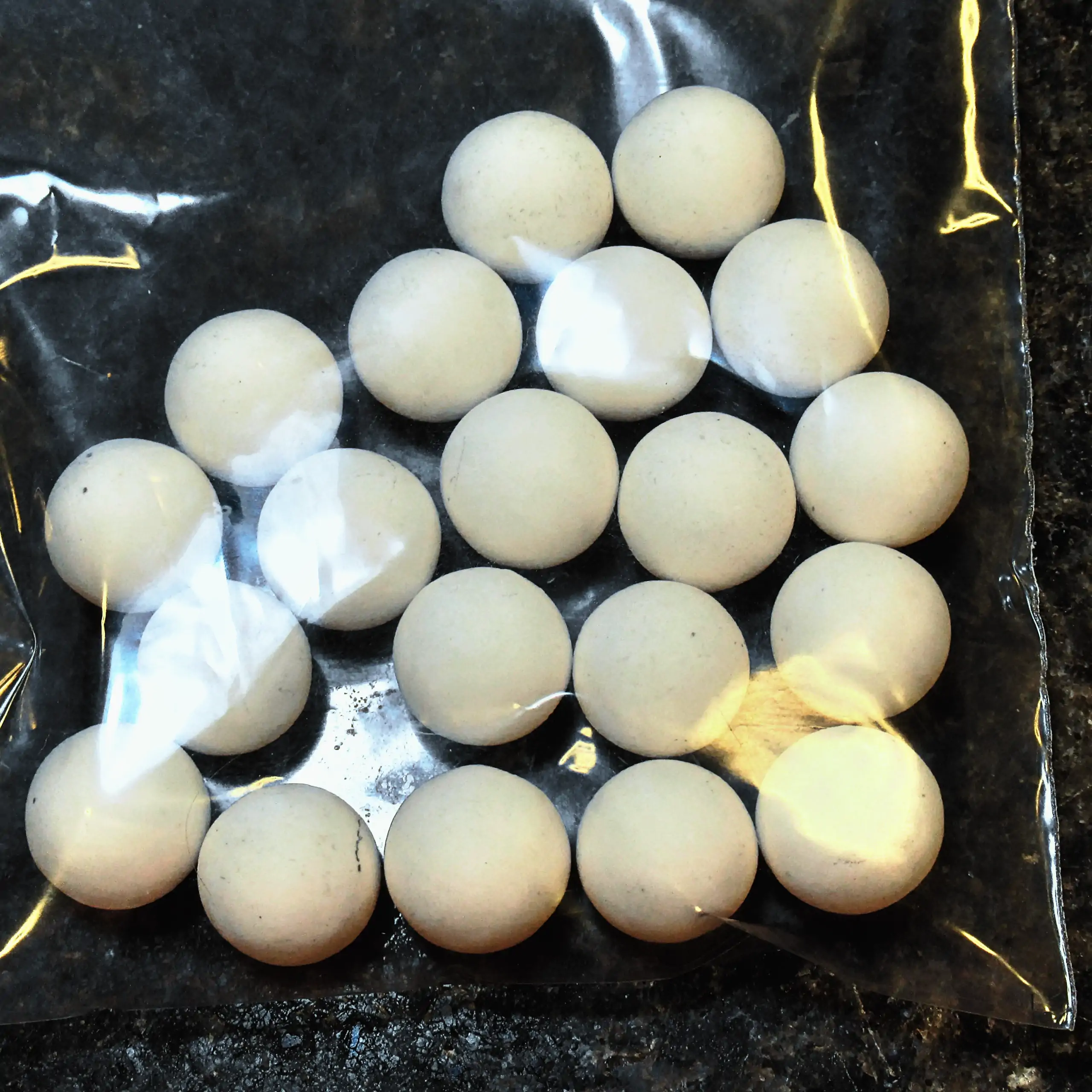 Ergänzende Ansicht der Körrie-Aktiv-Perlen (Keramik) / Körrie KAP S: Eine Gruppe leuchtend weißer, matter, runder Keramikperlen aus Aluminiumoxid in einer durchsichtigen Plastikverpackung, verstreut auf einer dunklen Oberfläche für eine reine, funktionale