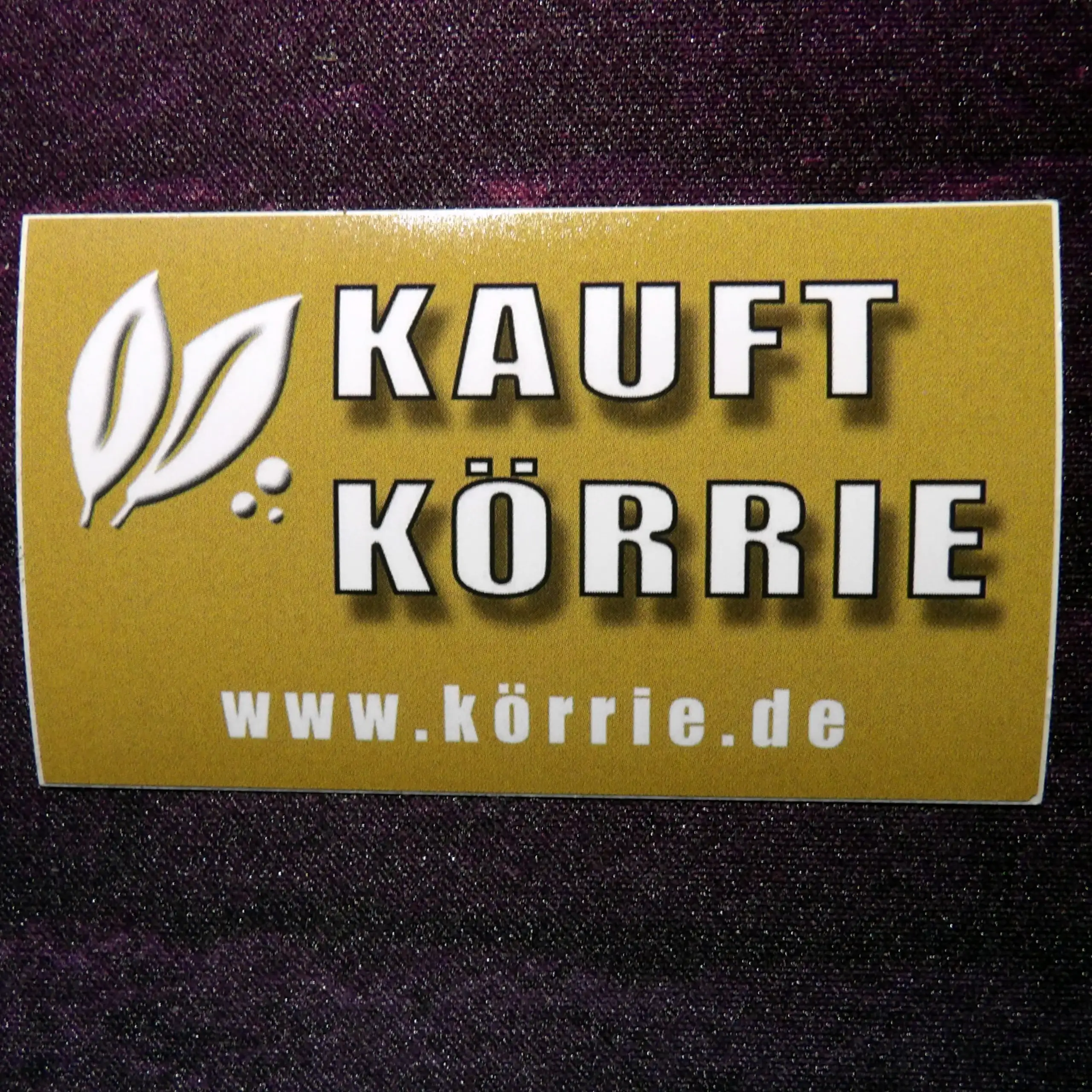 Originalaufkleber (Körrie KFA) "Kauft Körrie" auf dem Trägermaterial: Eine goldene, rechteckige Abbildung des Support-Aufklebers mit weißem Blatt-Logo, dem prominenten Claim "KAUFT KÖRRIE" in fetter Typografie und der Website www.körrie.de, die den Marken