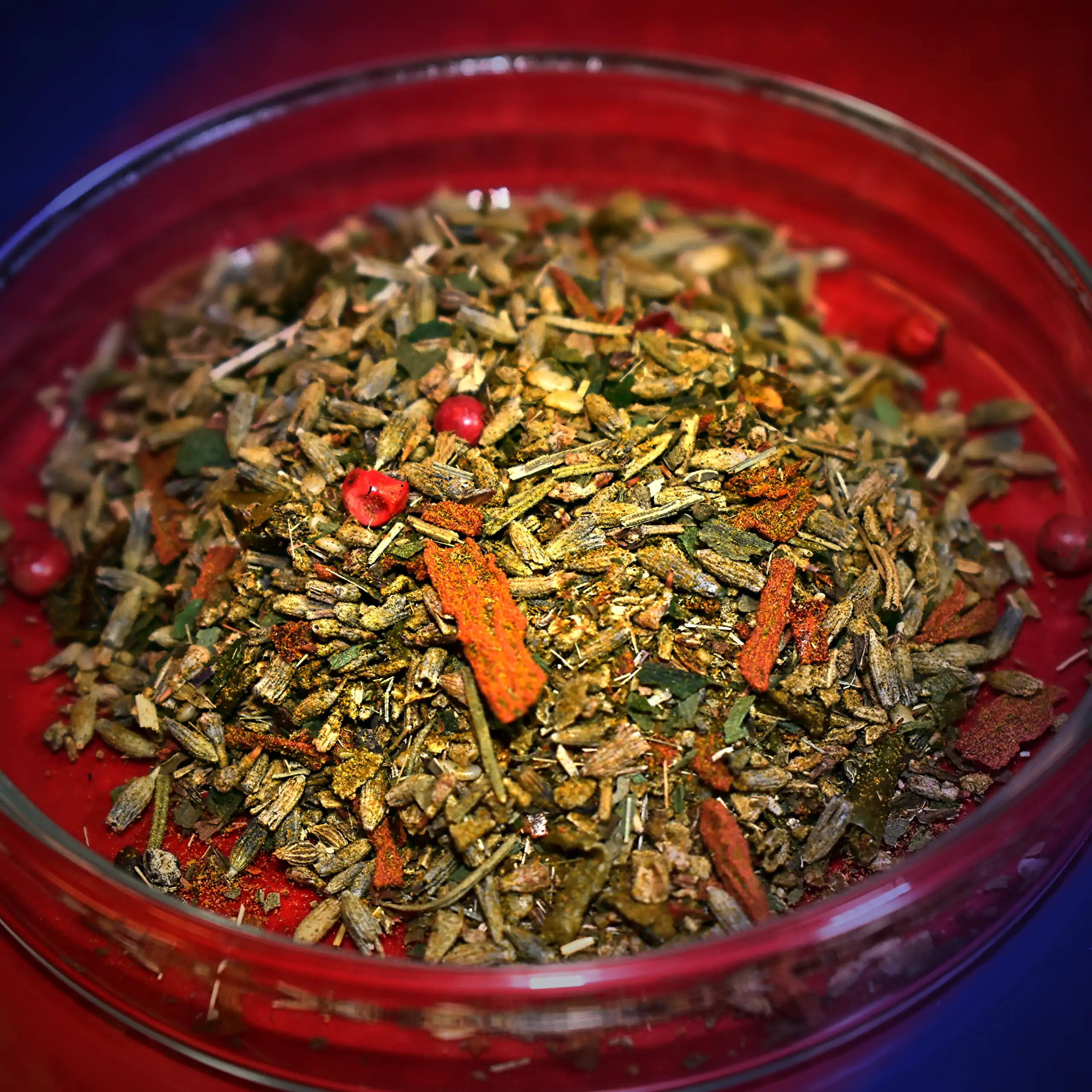 Ergänzende Ansicht der Körrie FLX Gewürzmischung: Eine farbenfrohe, grob geflockte Kräuter- und Gewürzmischung mit in einer transparenten Glasschale auf rotem Hintergrund mitwarmblauem Rahmen vignettiert, die natürlich flockige Textur und Vielfalt der "Pu