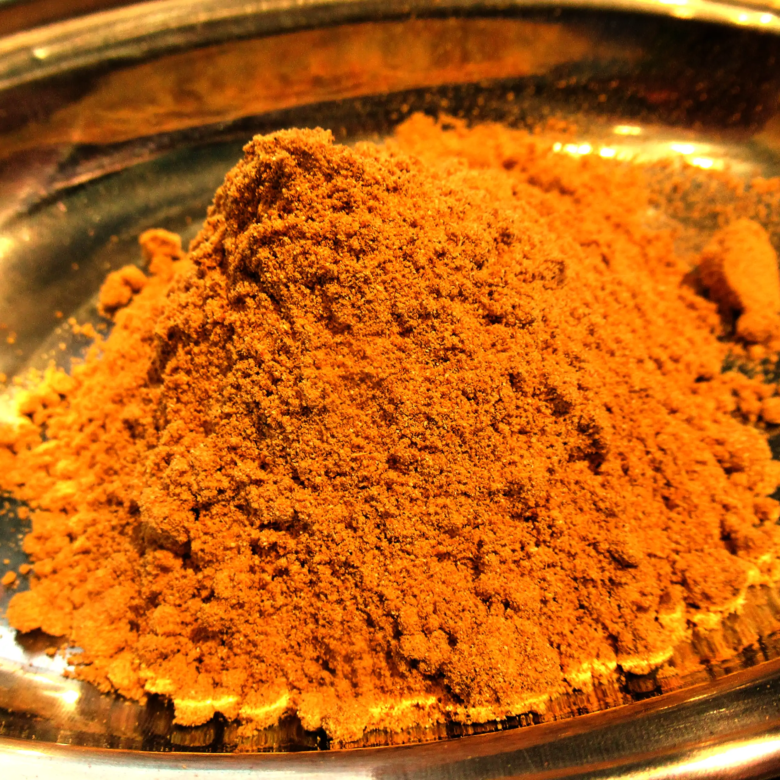 Ergänzende Ansicht der Körrie M25 Gewürzmischung: Ein Haufen leuchtend orange-goldener, fein gemahlener vollmundiger Würzmischung in einer metallischen Schale vor dunklem Hintergrund, der die intensive, aromatische Textur und warmgoldene Nuance betont, fü