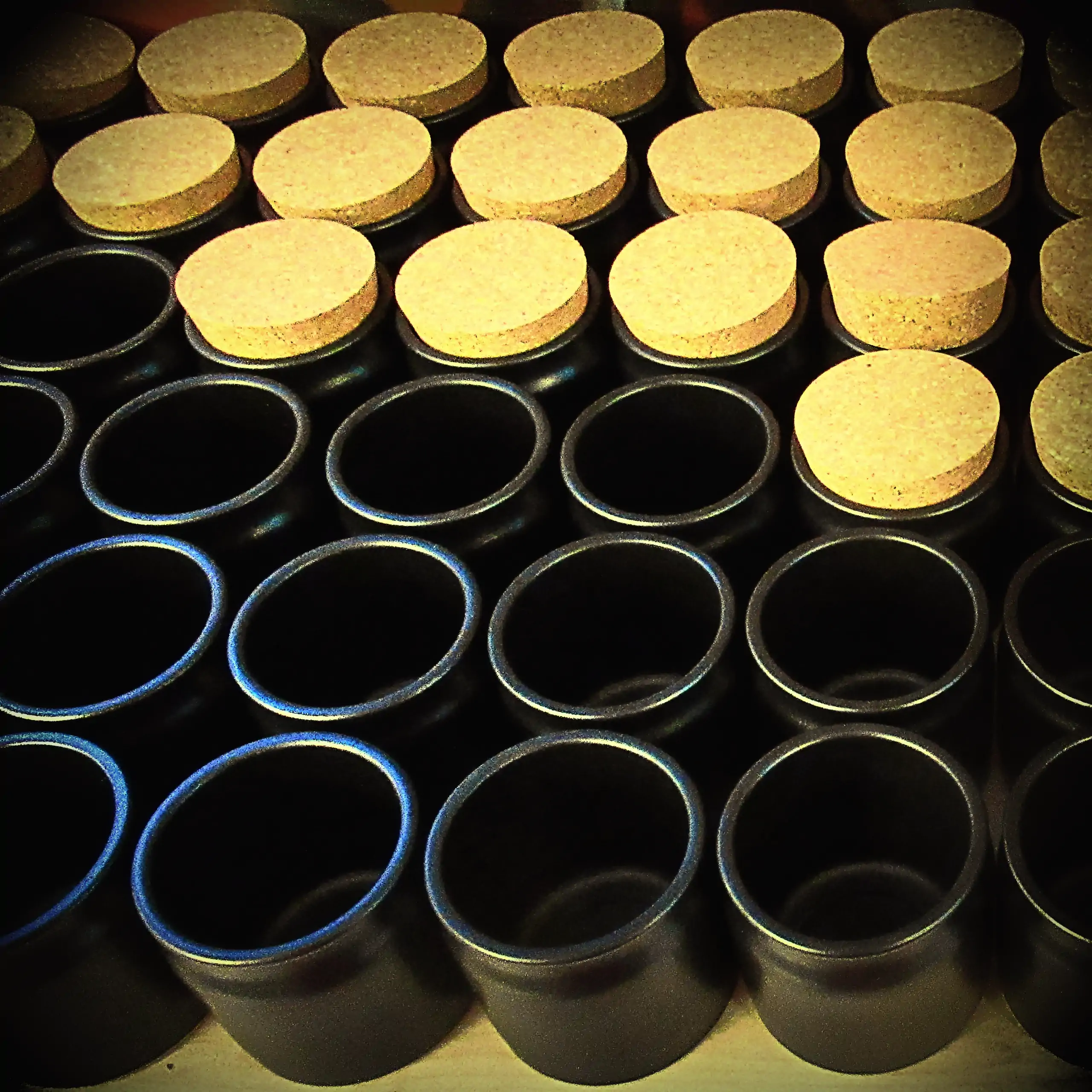 Ergänzende Ansicht der Körrie-Keramik-Töpfchen (Körrie KKT): Eine Reihe von eleganten, mattschwarzen Keramikgefäßen mit natürlichen Korkverschlüssen, geschlossen und offen in ordentlichen Reihen präsentiert auf einer gepolsterten Grundplatte für einen übe